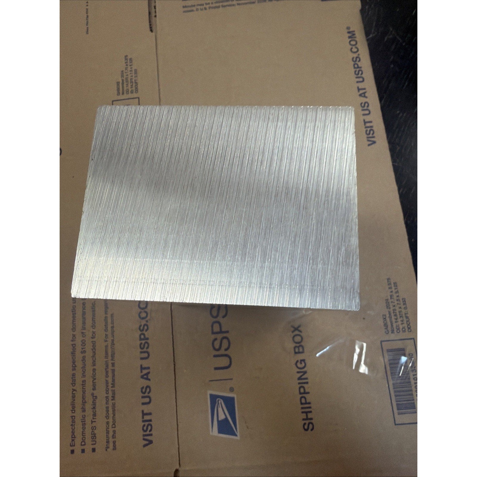 Rectangular metal plate with a fine vertical brushed texture, resting atop a USPS cardboard shipping box with visible text and logo.