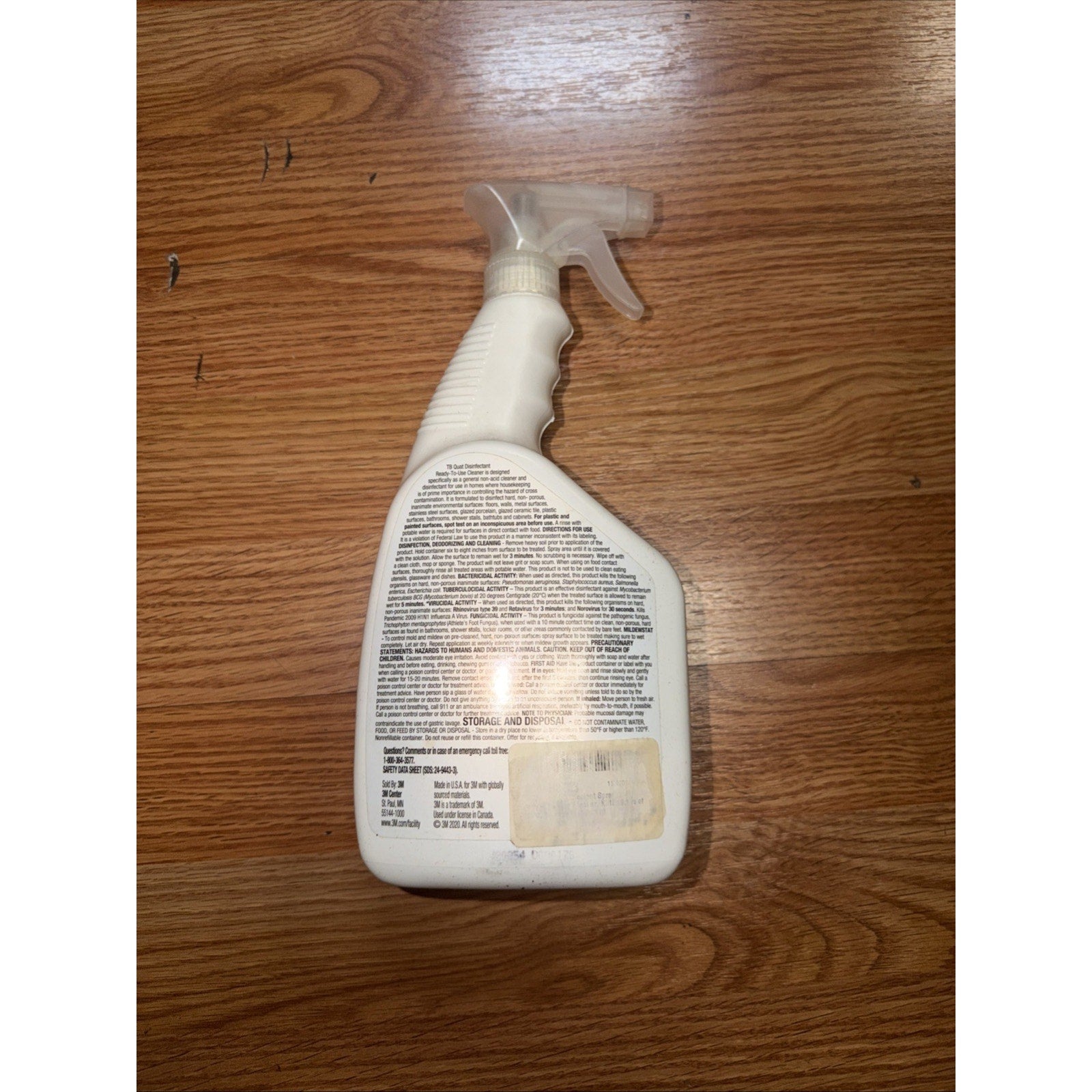 White plastic spray bottle with a clear trigger nozzle, showing detailed product information text on the back label, placed on a light brown wooden surface.
