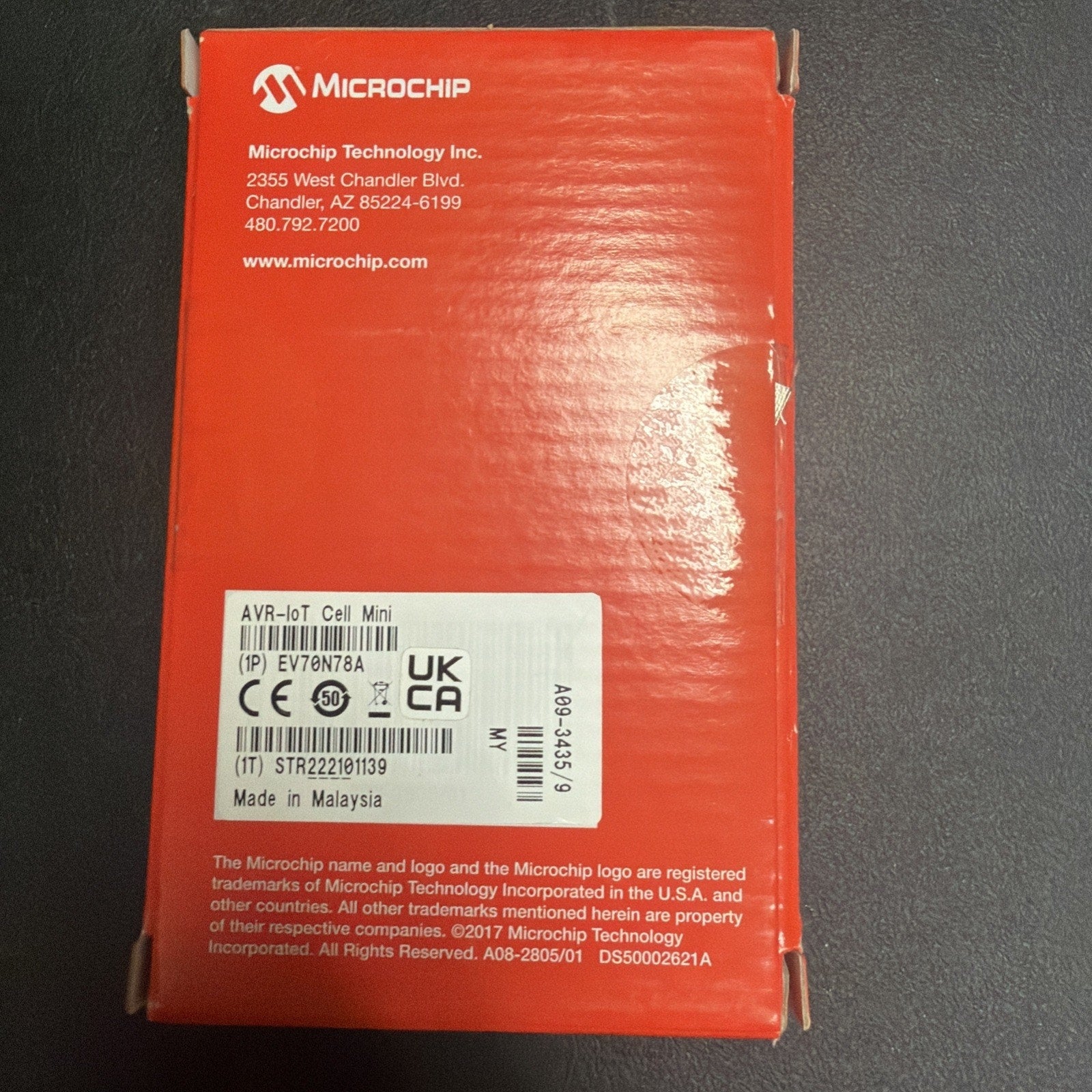 Red rectangular product box from Microchip Technology with company contact details, certification labels, and a white sticker indicating the model and manufacturing origin in Malaysia.