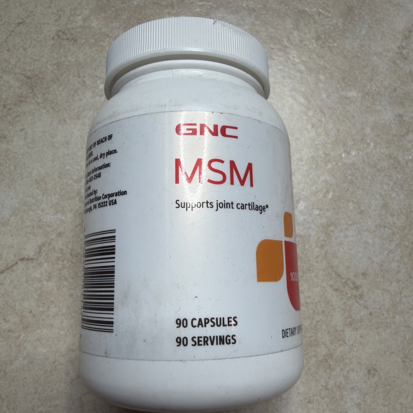 White plastic bottle of GNC MSM dietary supplement containing 90 capsules, labeled to support joint cartilage, with a barcode visible on the side against a gray textured background.