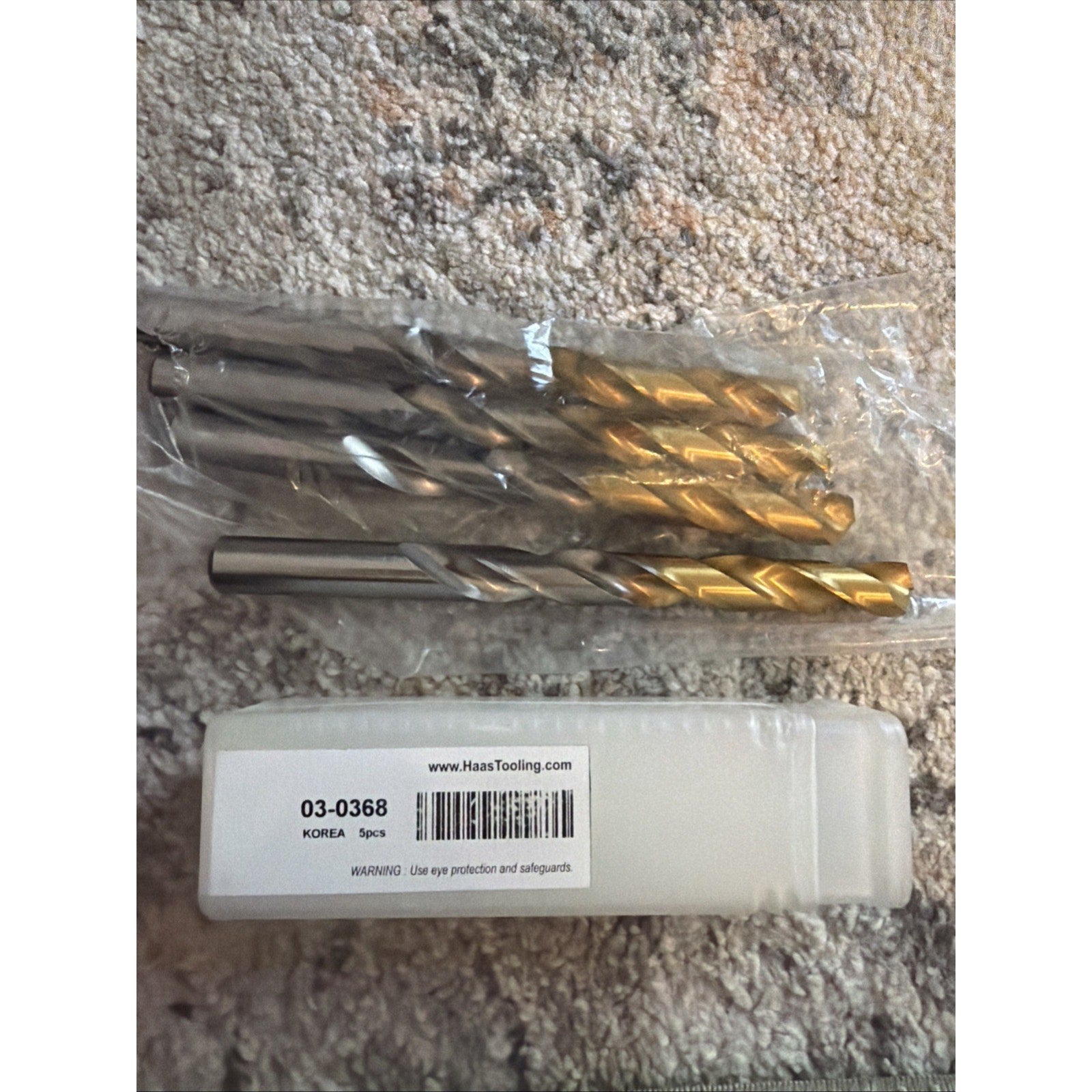 Five silver-colored metal drill bits with twist flutes arranged on a textured surface, partially inside a clear plastic sleeve, next to a white labeled plastic case.