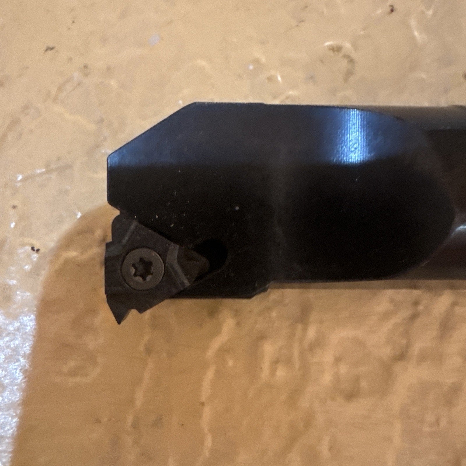 Close-up of a black metal cutting tool tip featuring a triangular carbide insert secured with a star-shaped screw, positioned against a textured beige surface.