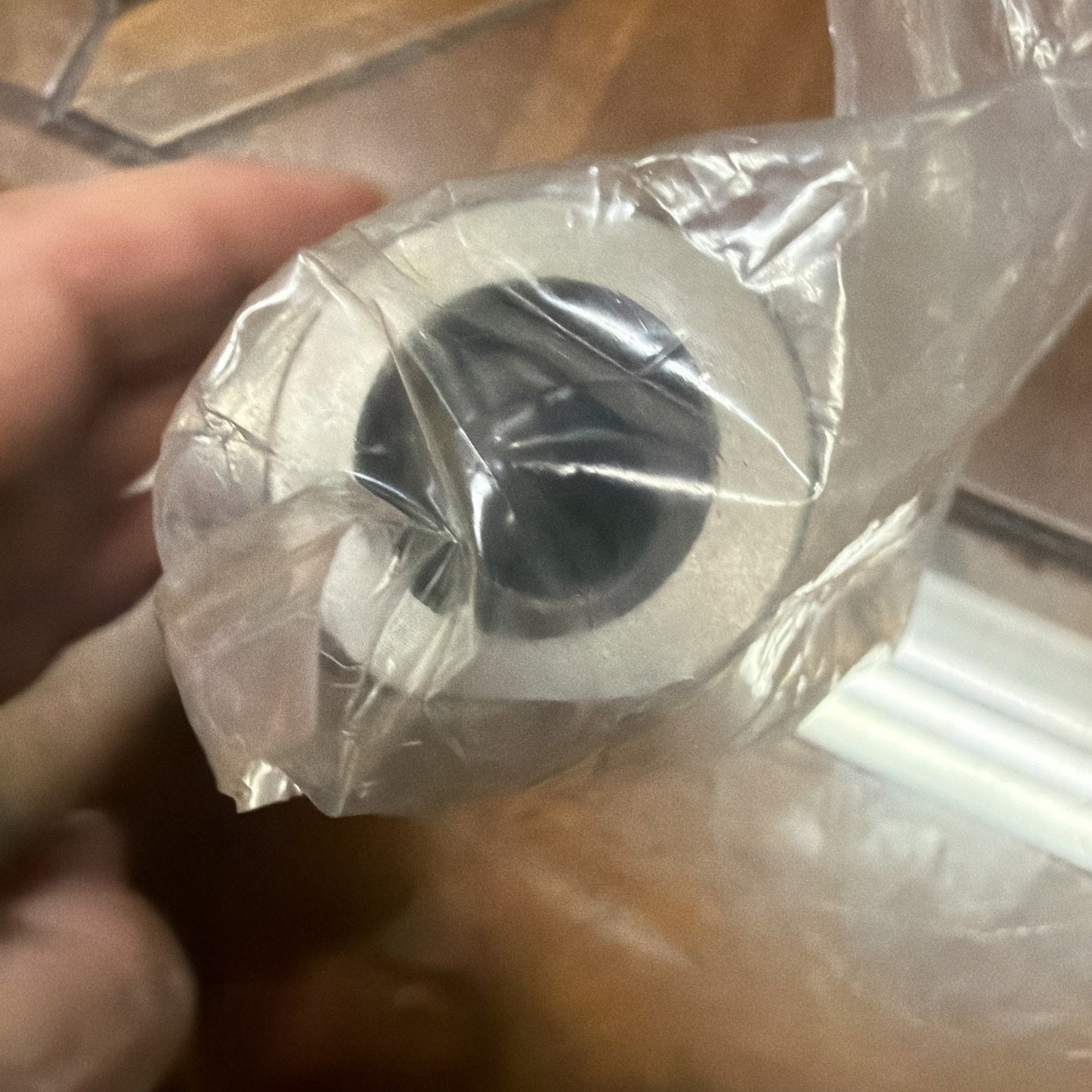 Hand holding a cylindrical metallic object wrapped in transparent plastic, showing a hollow center and smooth outer surface against a tiled floor background.