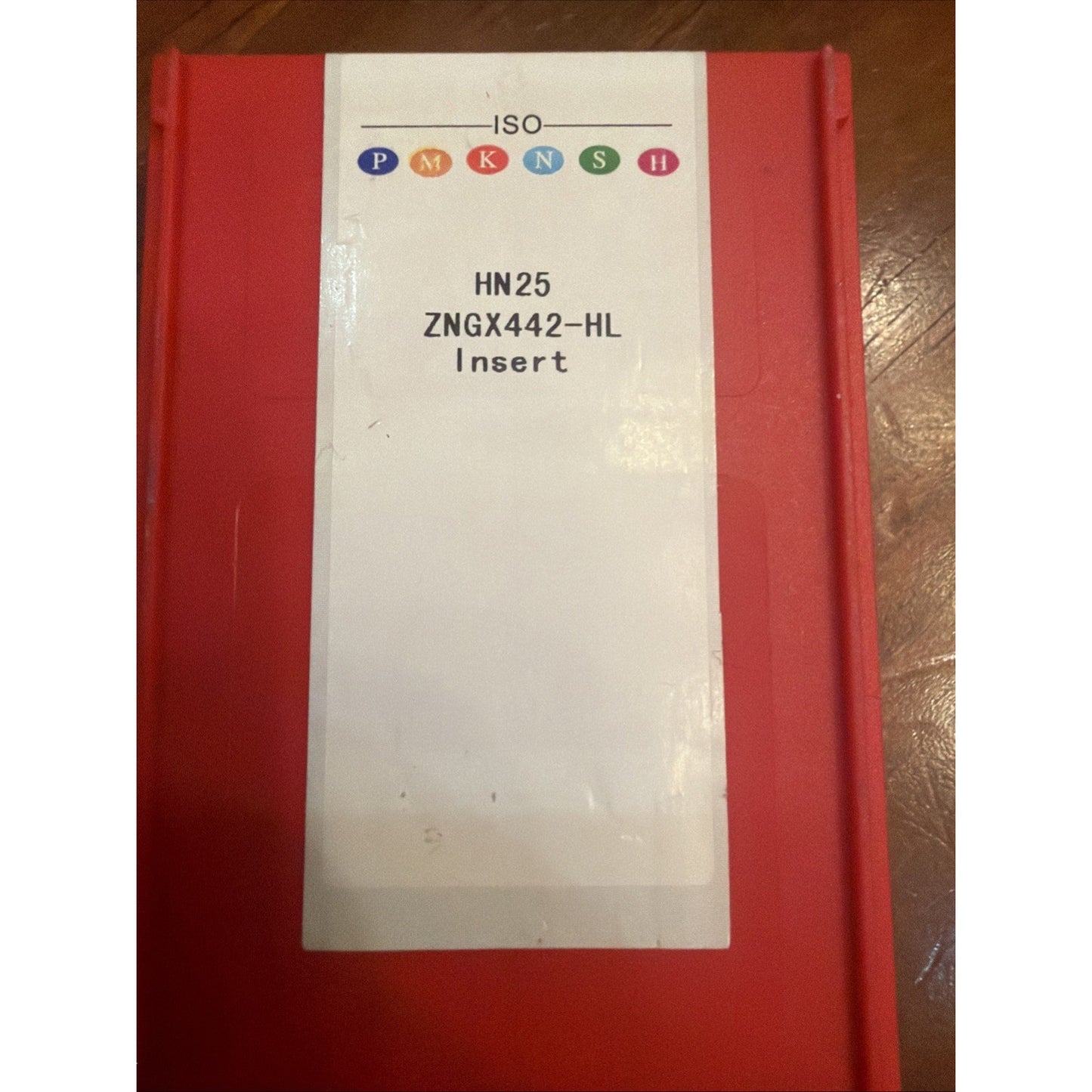 Red plastic container with a white label displaying ISO classification symbols and text indicating "HN25 ZNGX442-HL Insert," placed on a wooden surface.