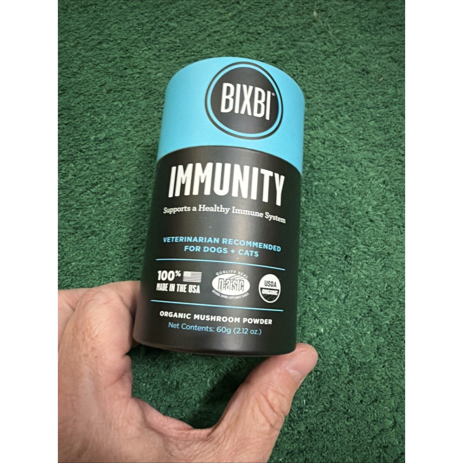Hand holding a cylindrical container of Bixbi Immunity organic mushroom powder for dogs and cats on a green textured background.