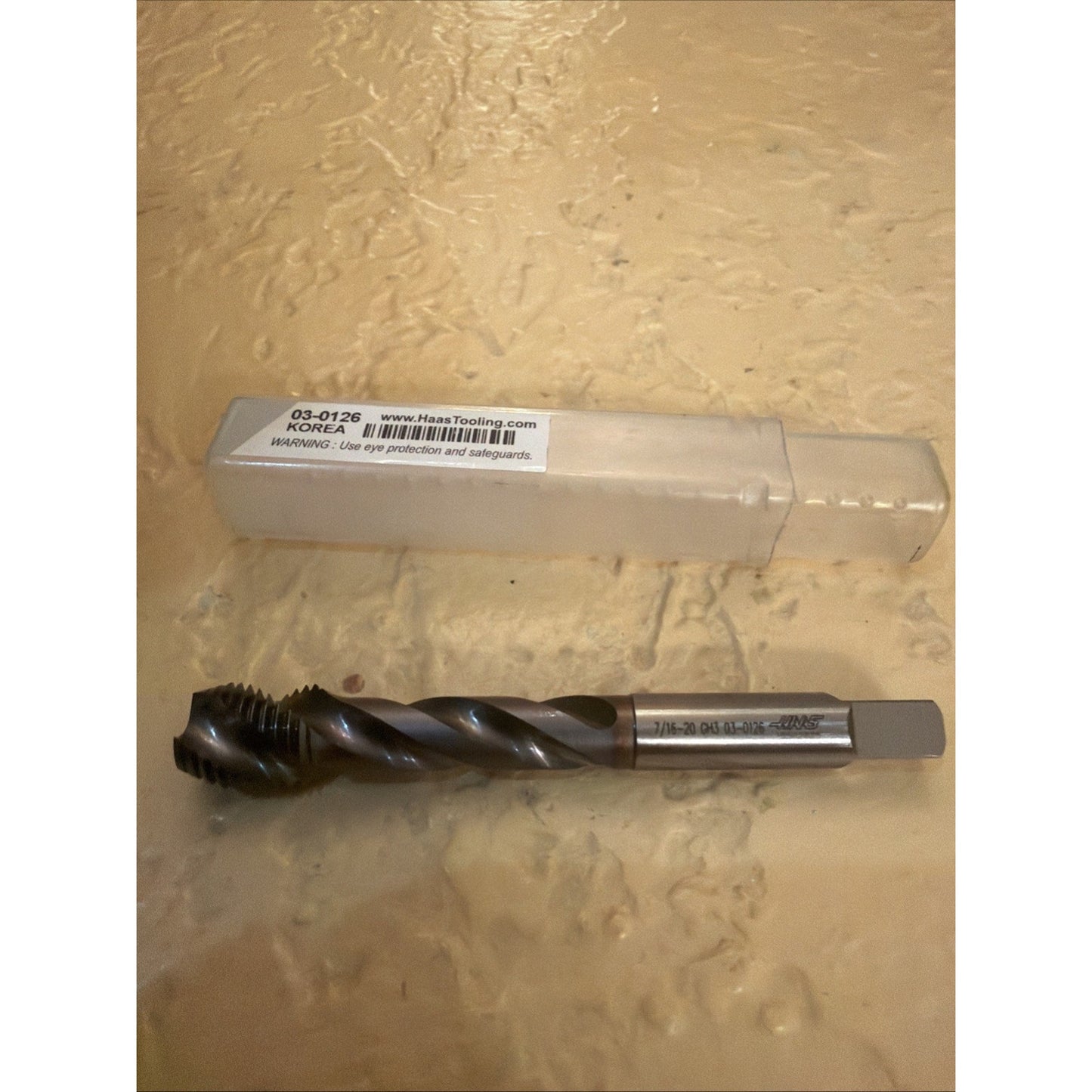 Single metal spiral threading tap with a square drive shank, positioned below a translucent plastic storage case with a barcode label and cautionary text on a textured beige surface.