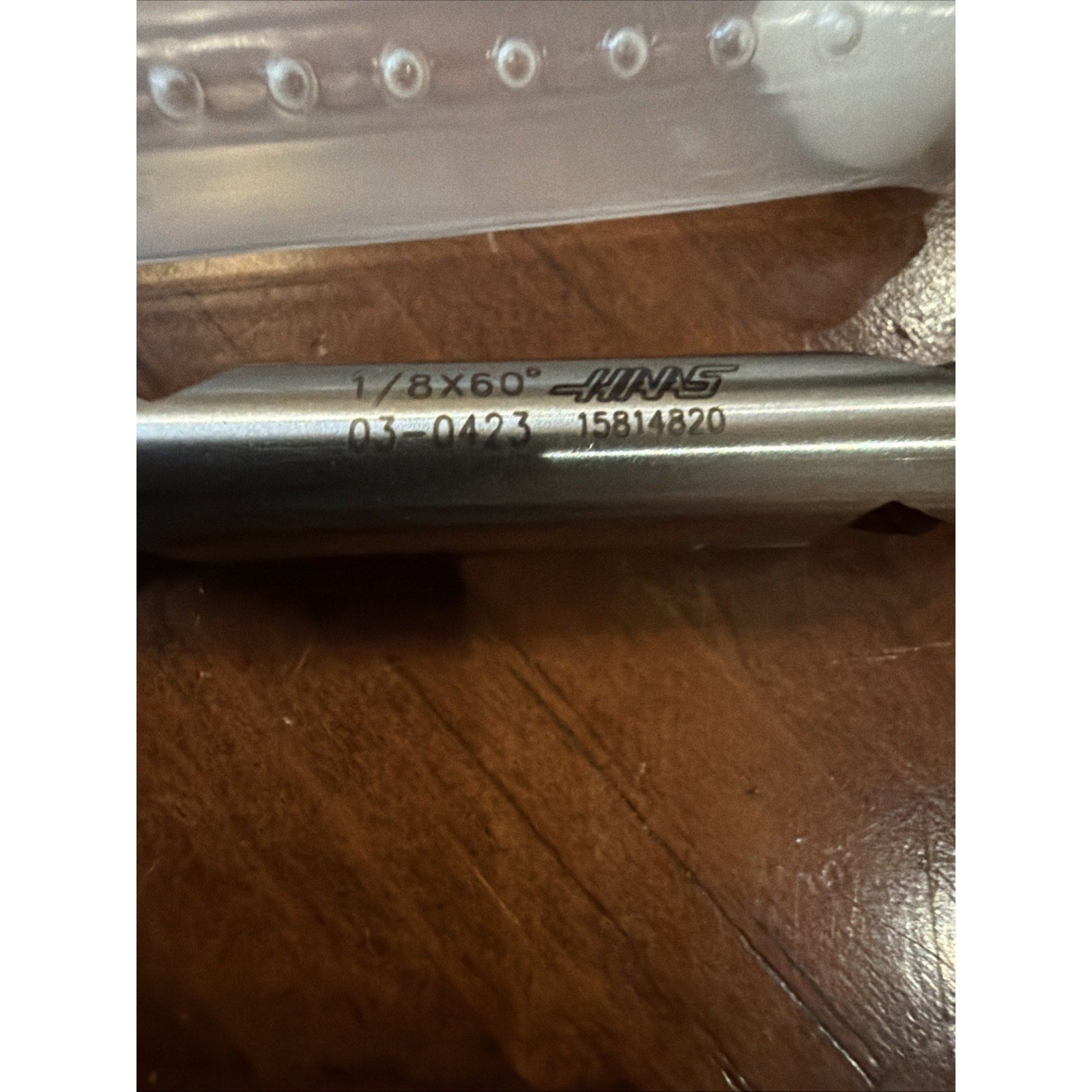 Close-up of a metal tool with etched markings "1/8X60°," the Haas logo, a product code, and serial number, resting on a wooden surface beneath a clear plastic case.