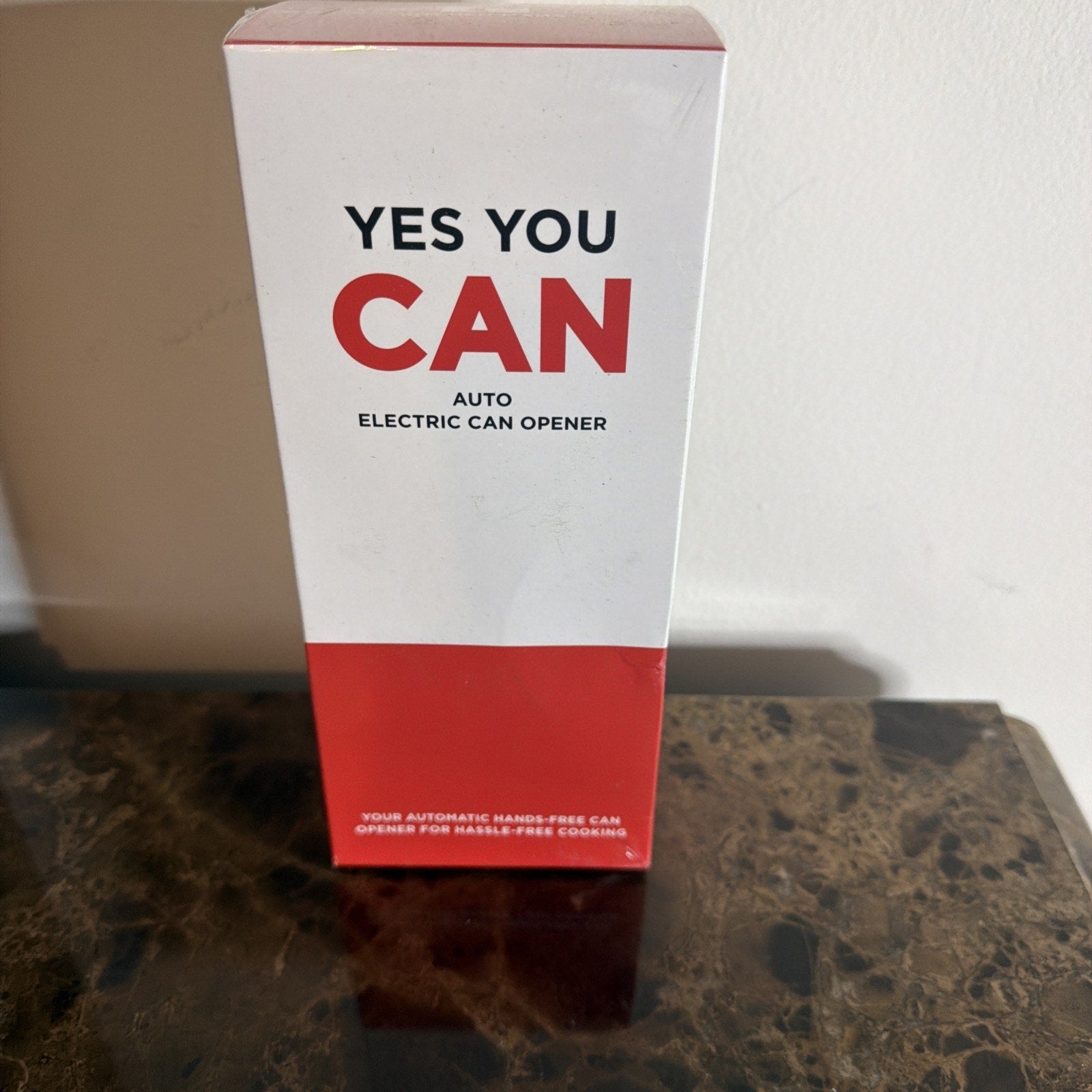 Tall white and red rectangular box standing on a dark marble surface with text reading "YES YOU CAN" and "Auto Electric Can Opener" on the front.