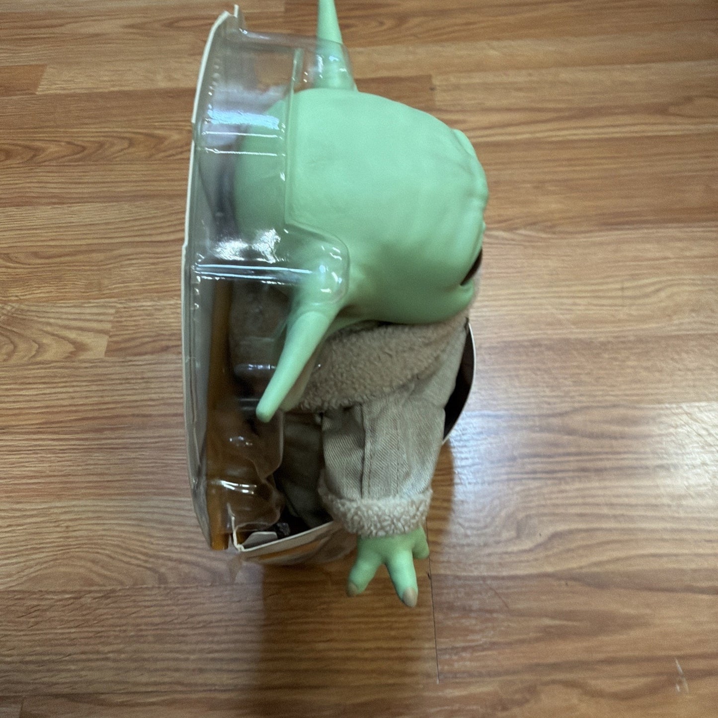 Side view of a green, alien-like figure with large ears wearing a beige jacket with a fleece collar, partially enclosed in clear plastic packaging on a wooden surface.