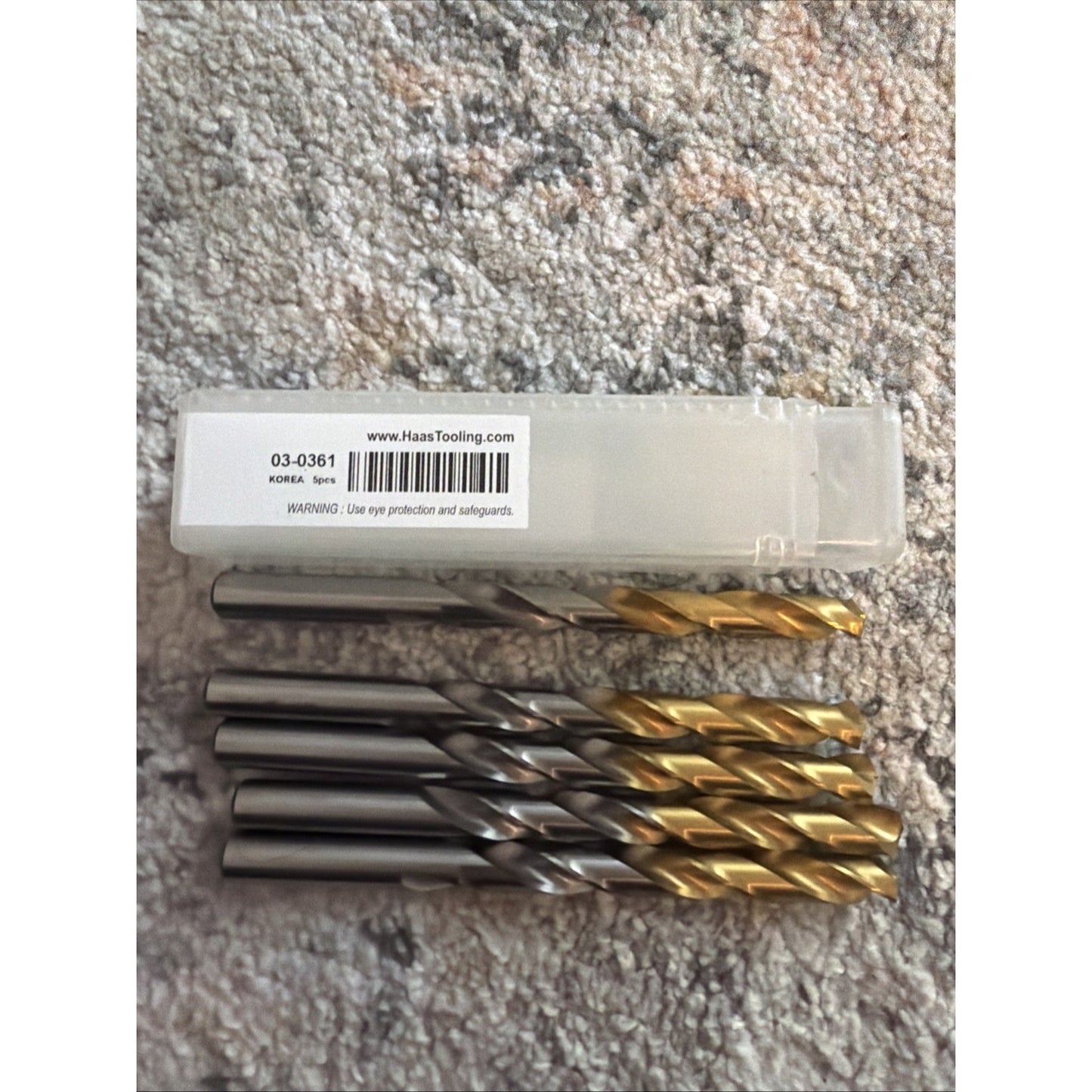 Set of five metal drill bits with silver and gold-colored coated spiral flutes, arranged in front of a clear plastic storage box with a white label, on a textured light carpet surface.