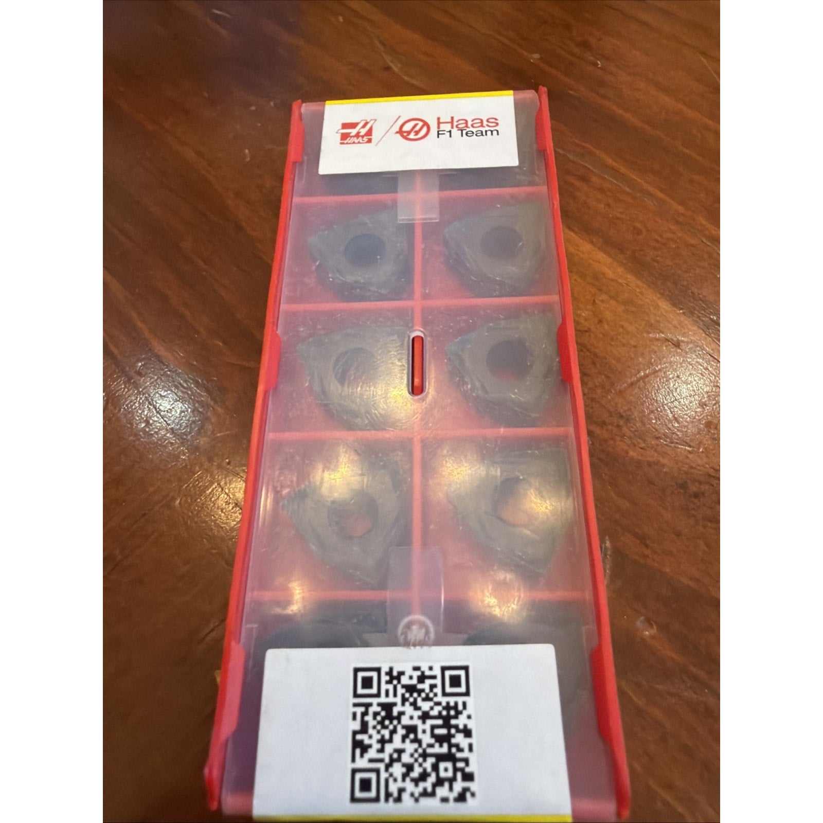 Red plastic case containing six triangular metal inserts with labeled Haas F1 Team logo and a QR code on a wooden surface.