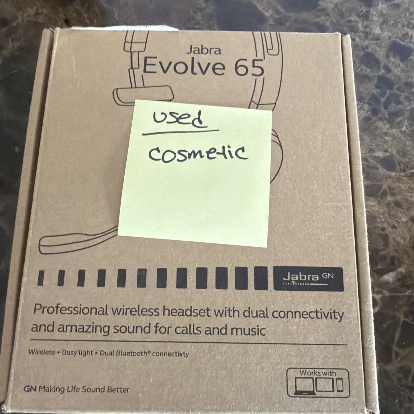 Brown cardboard box packaging for a Jabra Evolve 65 wireless headset showing product branding, features of dual Bluetooth connectivity, sound quality for calls and music, and a yellow sticky note labeled "used cosmetic" placed on top.