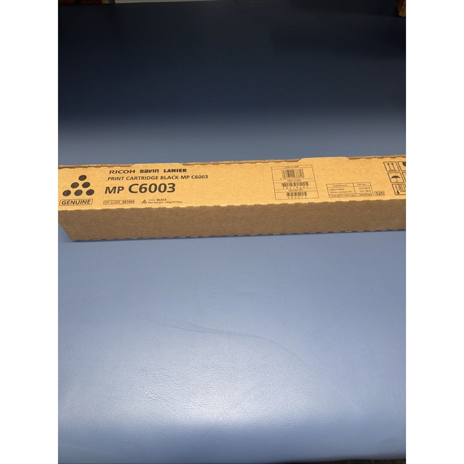 Brown cardboard box packaging for a genuine Ricoh MP C6003 laser print cartridge, featuring black printed text and manufacturing details on a plain blue surface background.