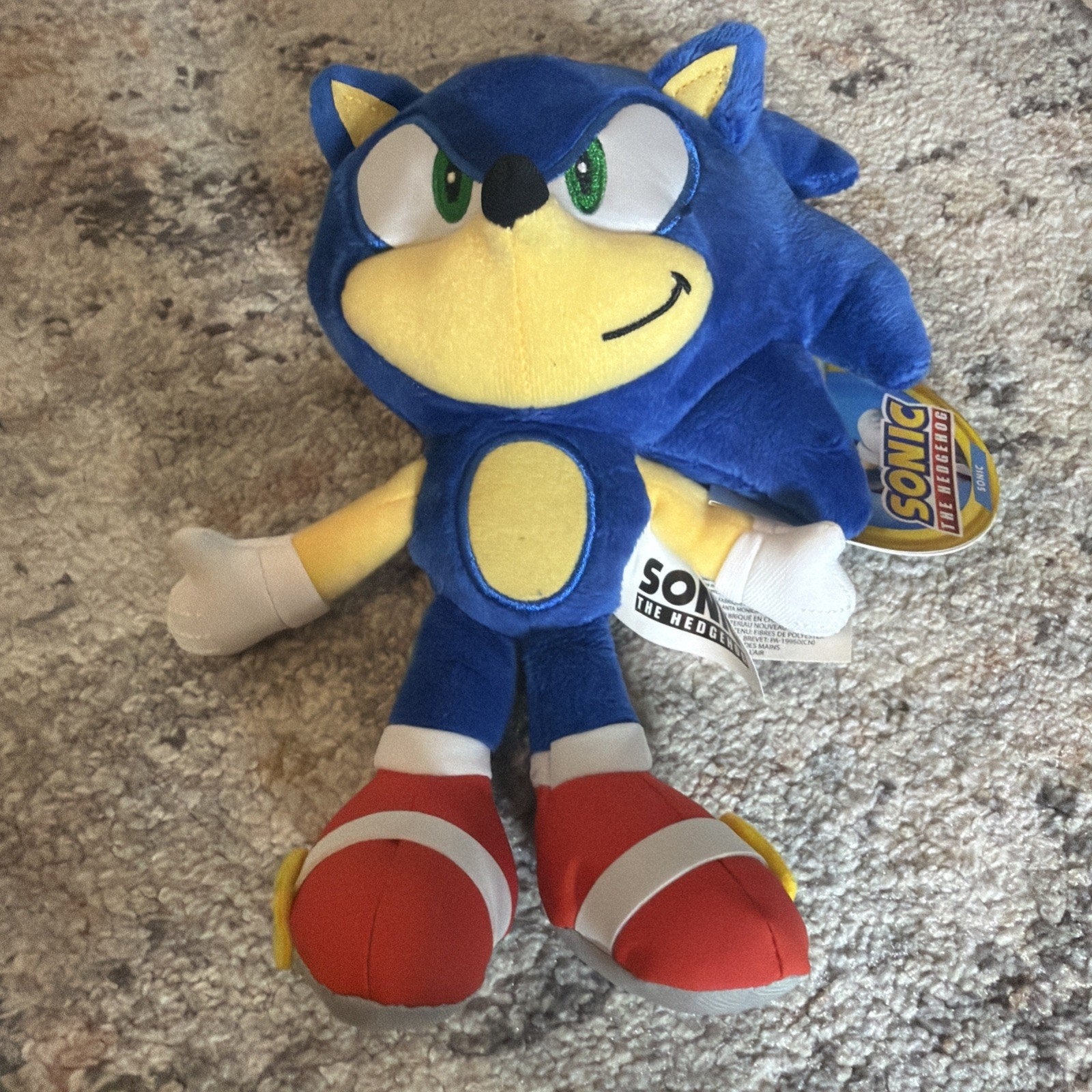 Plush toy of a blue character with green eyes, yellow arms, white gloves, and red shoes with white straps, lying on a textured carpet with attached product tags.