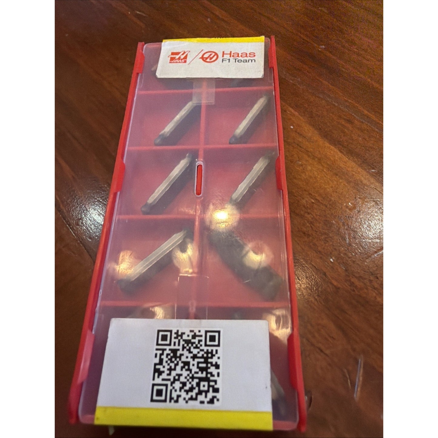 Red plastic container with a translucent lid holding eight metal cutting inserts arranged in individual compartments, featuring Haas F1 Team branding and a QR code label on a wooden surface.