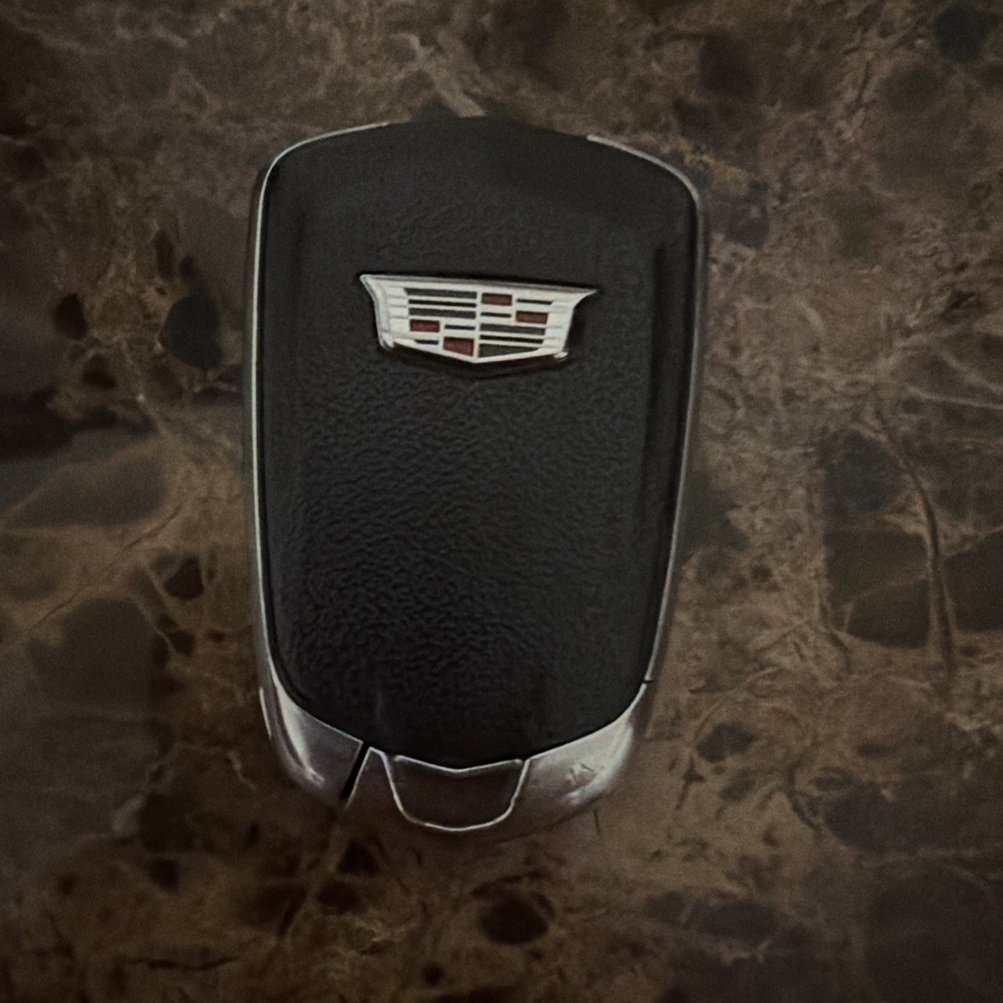 Black car key fob with a silver Cadillac emblem on the front, resting on a dark brown marbled surface.