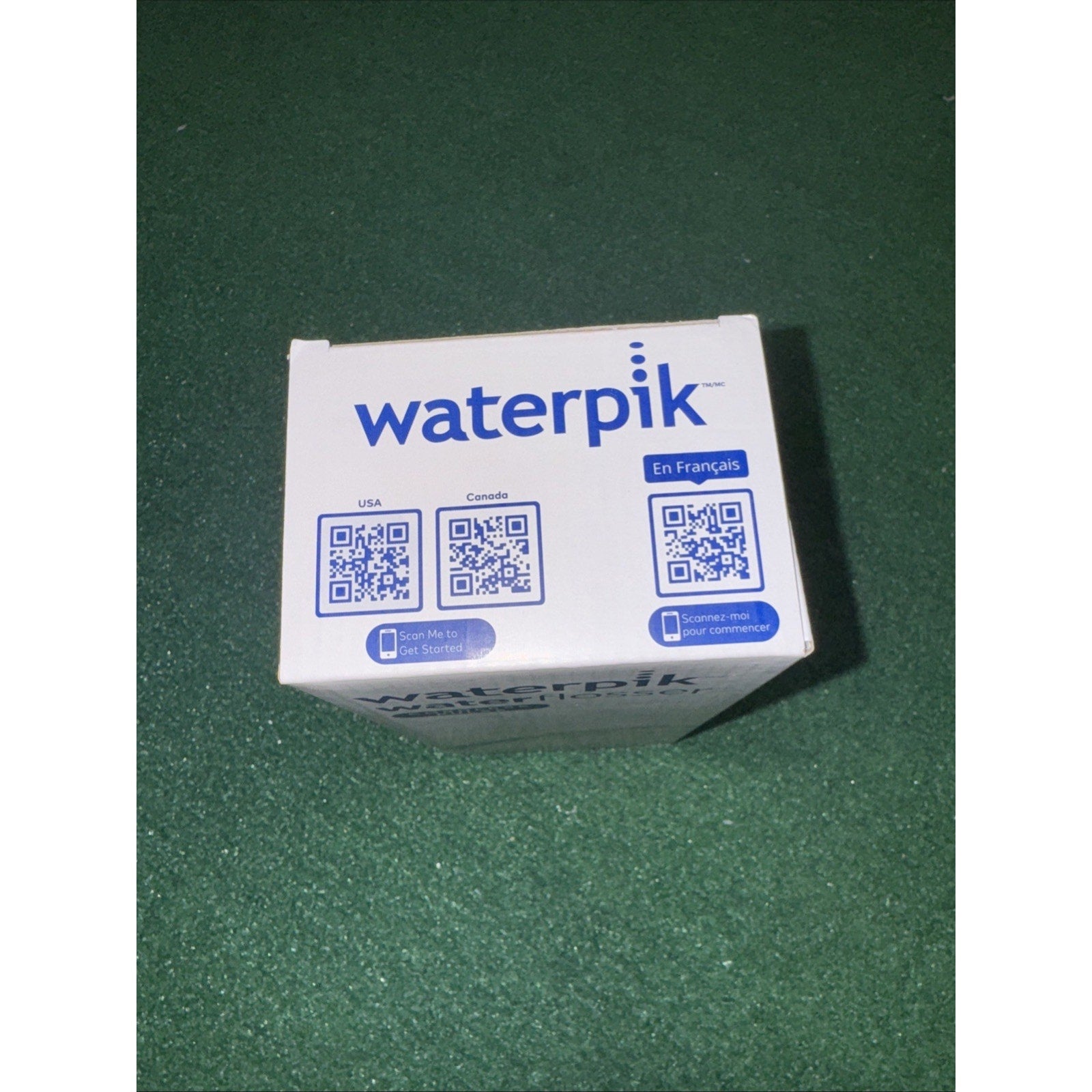 Top view of Waterpik product box showing blue logo and QR codes on white packaging against green background
