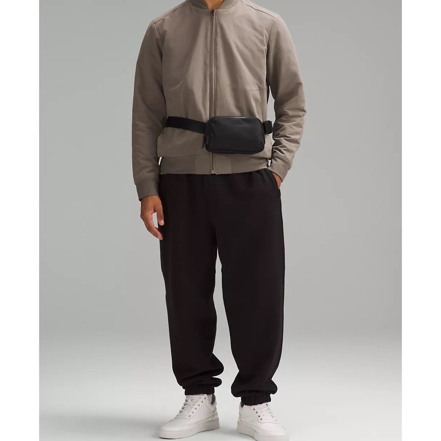 Person wearing a taupe zip-up jacket and black jogger pants paired with white sneakers, accessorized with a small black waist bag against a plain gray background.