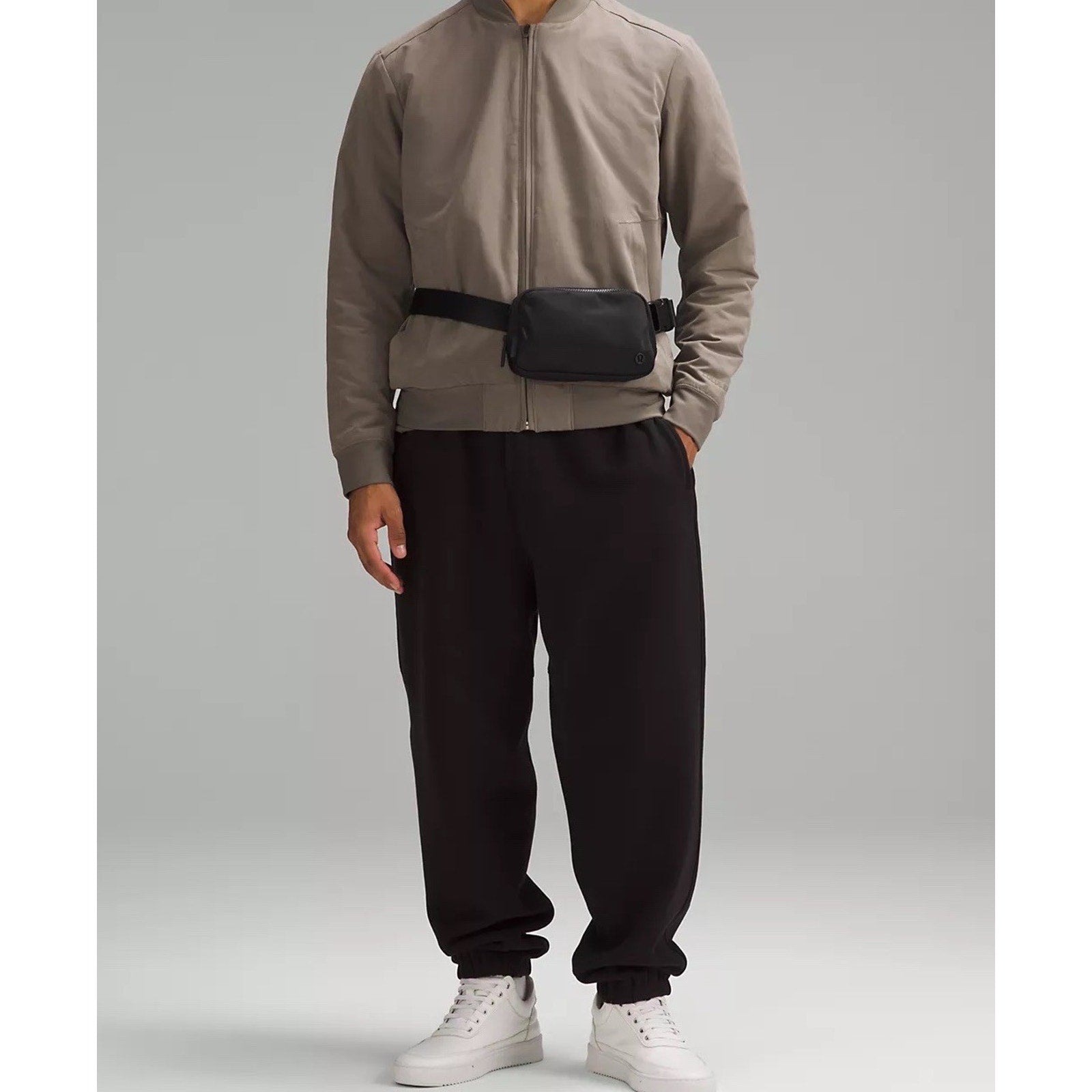 Person wearing a taupe zip-up jacket and black jogger pants paired with white sneakers, accessorized with a small black waist bag against a plain gray background.