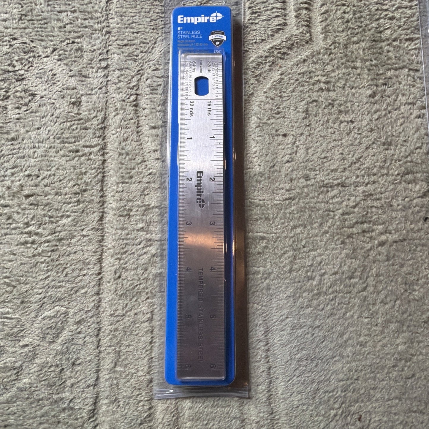Steel ruler with inch and centimeter markings packaged in clear plastic with a blue Empire-branded cardboard backing, placed on a textured beige carpet.