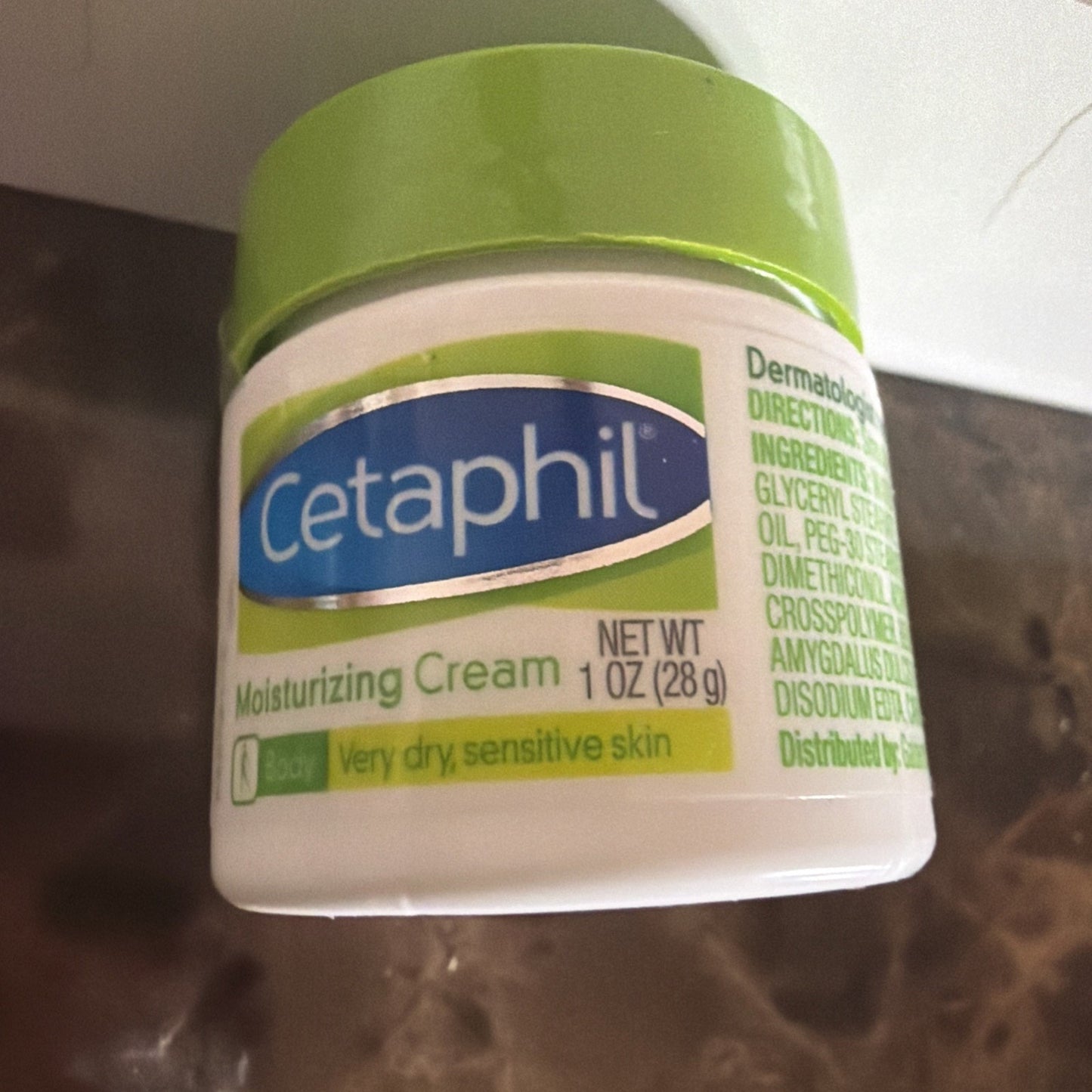 White plastic jar of Cetaphil Moisturizing Cream with a light green lid, labeled for very dry, sensitive skin, placed on a brown marble surface near a white wall.