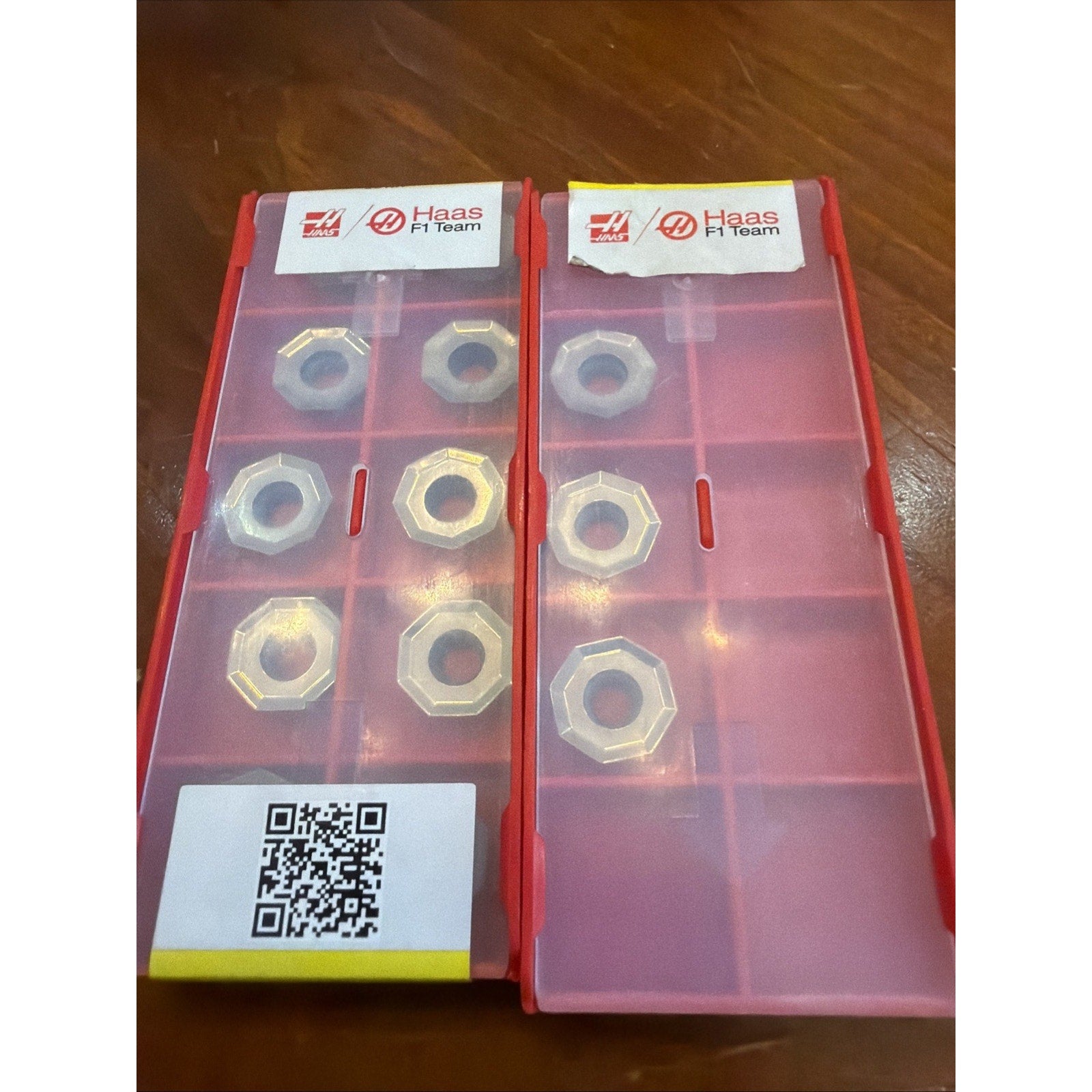 Two red plastic compartmentalized containers with translucent lids, each holding hexagonal metal inserts, featuring Haas F1 Team branding and a QR code on one container, placed on a wooden surface.