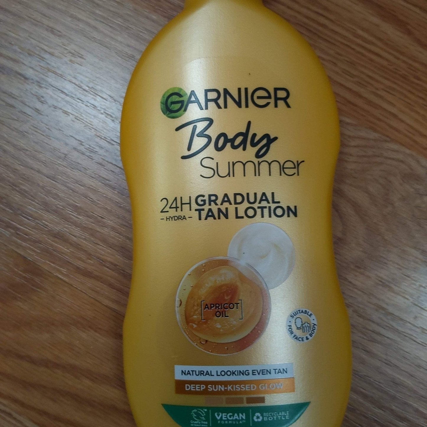 Golden yellow Garnier Body Summer 24H gradual tan lotion bottle with apricot oil, showing natural tan and deep sun-kissed glow claims on a wooden surface.