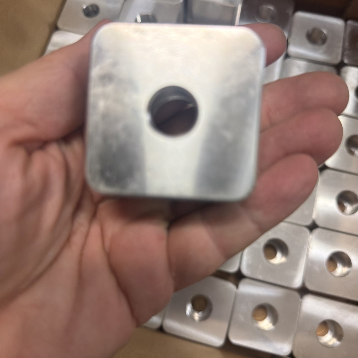 Hand holding a small, square metal block with a centered circular hole, with multiple identical metal blocks arranged in the background.