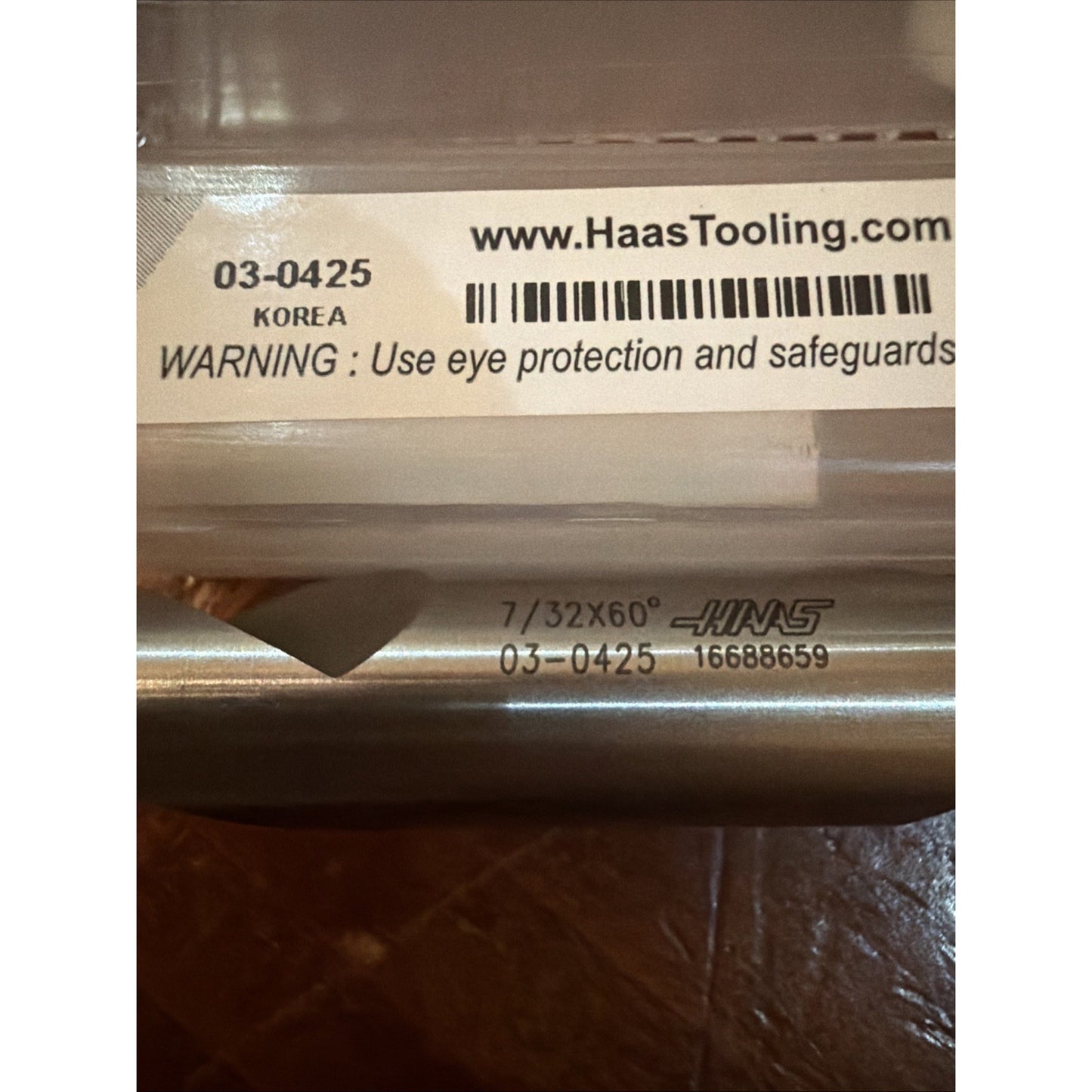 Close-up of a metal cutting tool with engraved text "7/32X60°," "HAAS," and serial number, resting near a clear plastic case label showing a barcode, website, and safety warning.