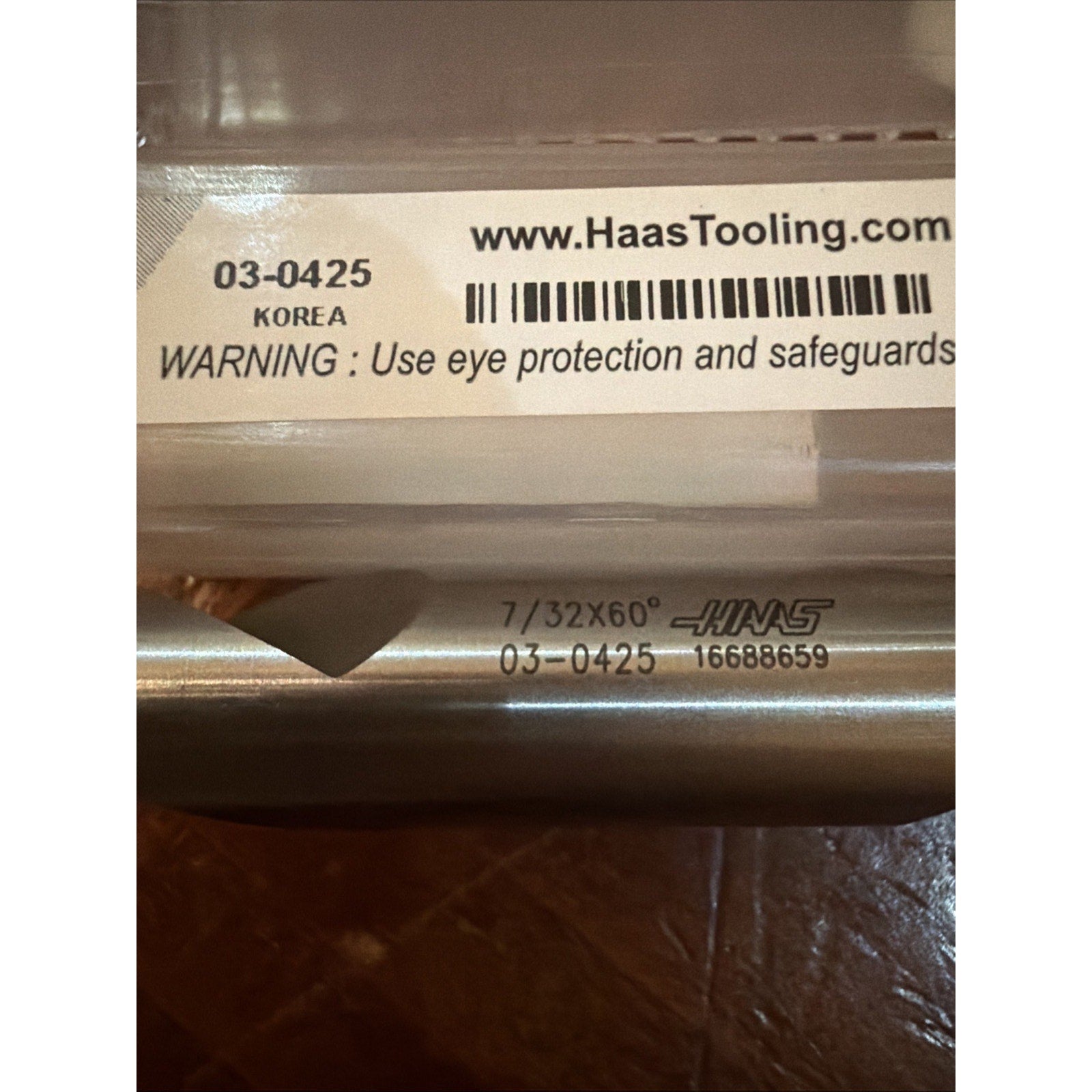 Close-up of a metal cutting tool with engraved text "7/32X60°," "HAAS," and serial number, resting near a clear plastic case label showing a barcode, website, and safety warning.