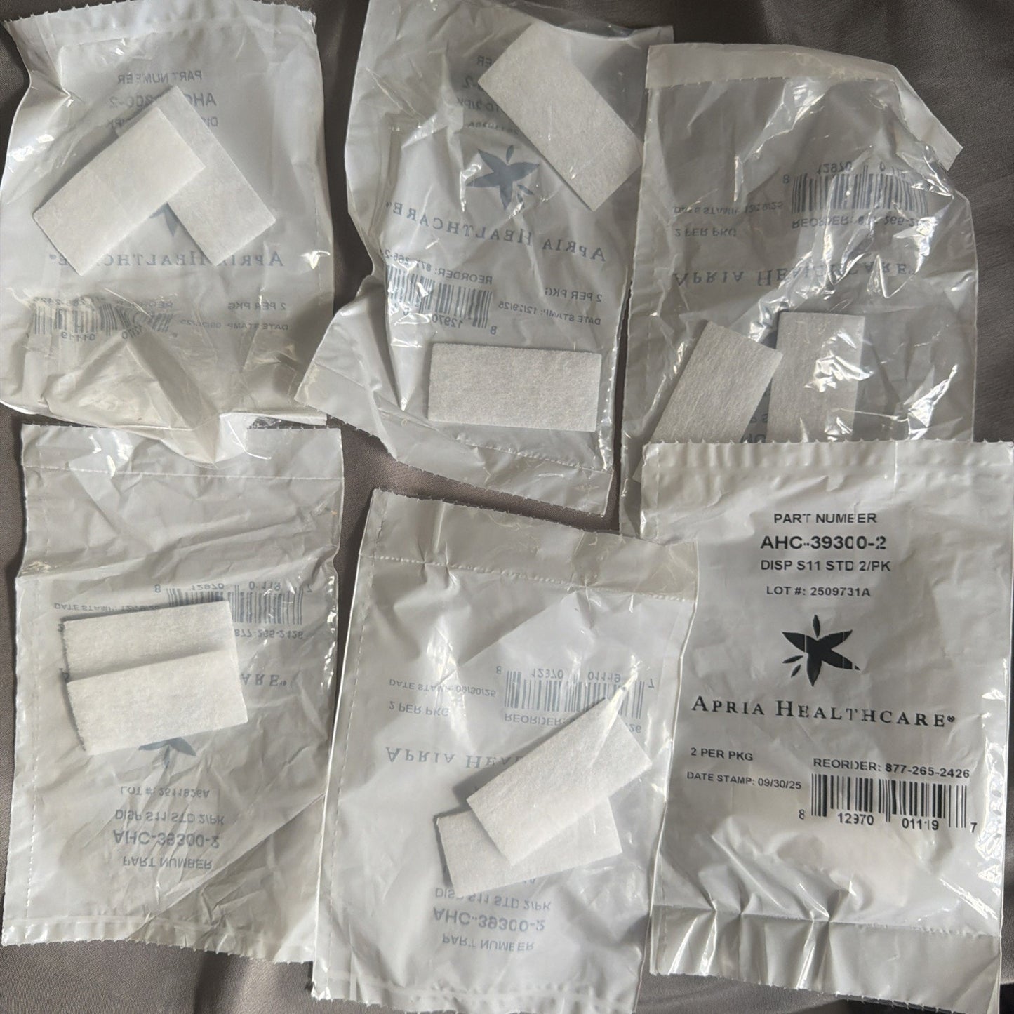 Six clear plastic packages containing small white fabric strips, with one package showing a label for Apria Healthcare featuring part number, batch information, and contact details.