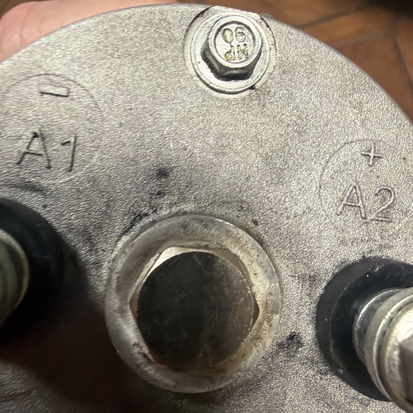 Close-up view of a metallic circular component with a central hexagonal bolt, two terminals labeled "A1" and "A2" with plus and minus symbols, and a small stamped number near a screw.