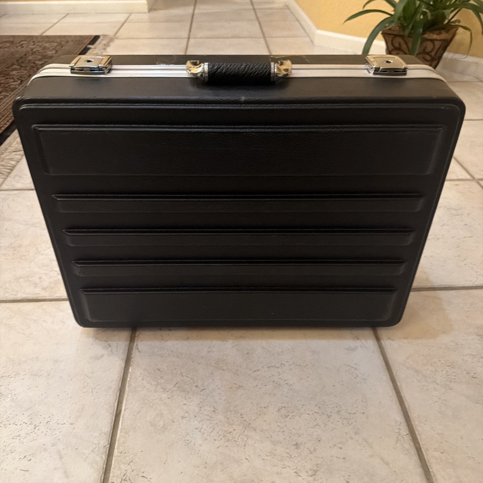 Black hard-sided briefcase with horizontal indent designs on the front, silver metal latches, and a textured black handle, placed on a tiled floor indoors.