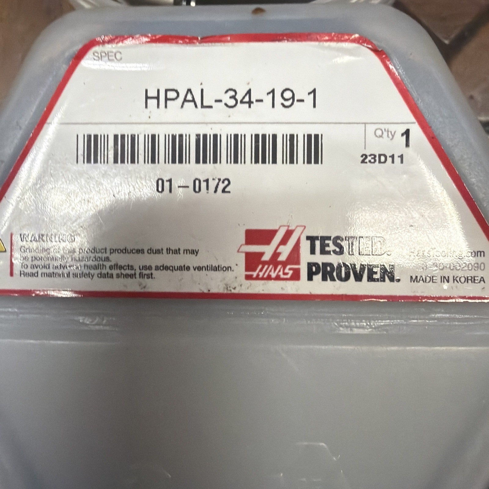 Rectangular white label with red border displaying product code "HPAL-34-19-1," barcode, quantity "1," and warning about dust hazards, attached to a gray plastic container.