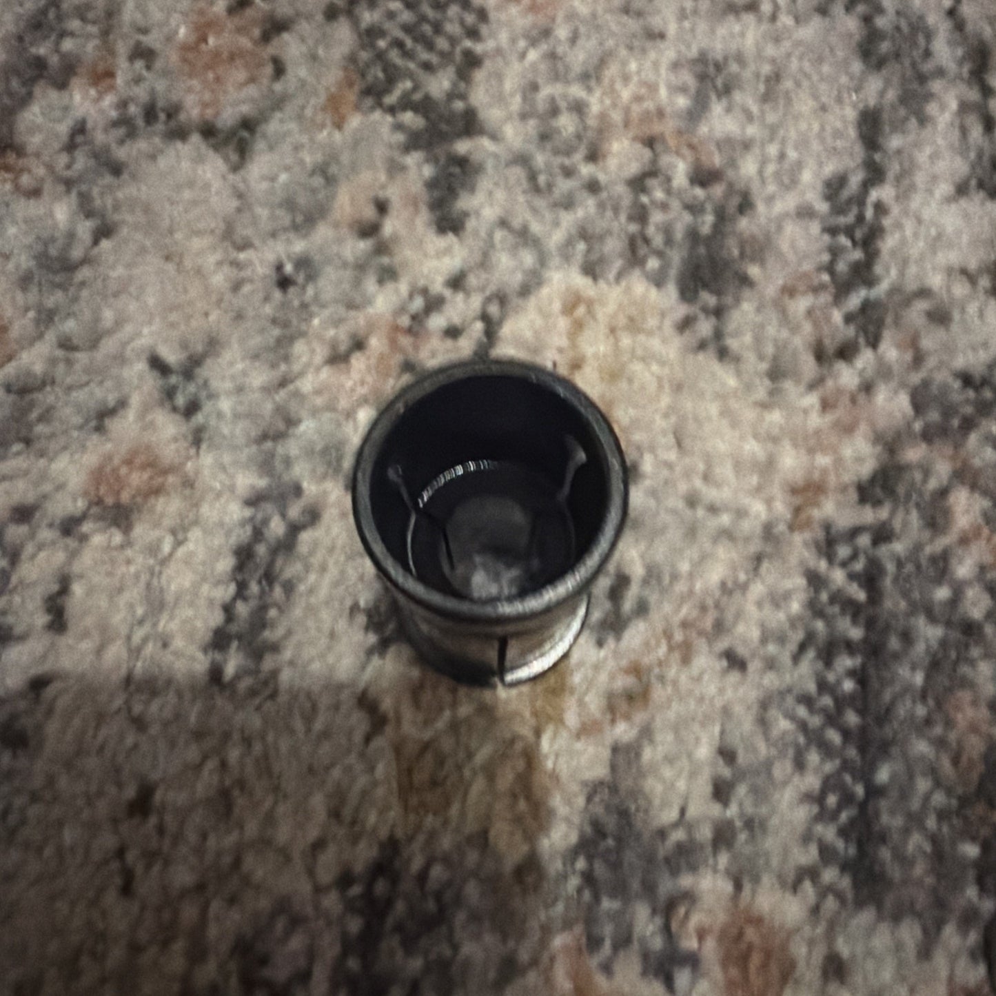 Small black cylindrical metal part with a hollow center, resting upright on a patterned carpet with muted earth tones.