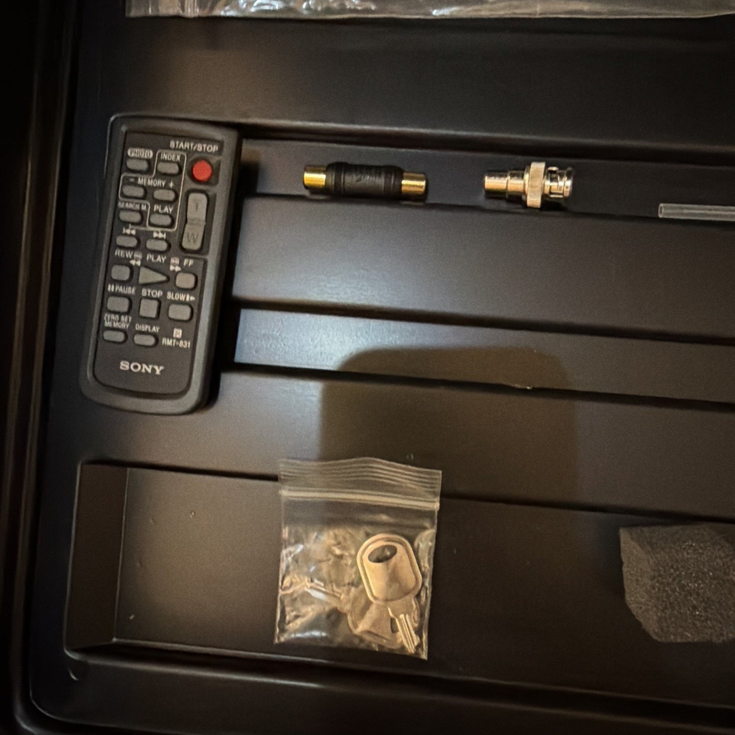 Black remote control with white buttons labeled Sony, a short RCA adapter cable, a metal connector with a screw lock, and a small plastic bag containing two keys arranged in a black foam-lined case.