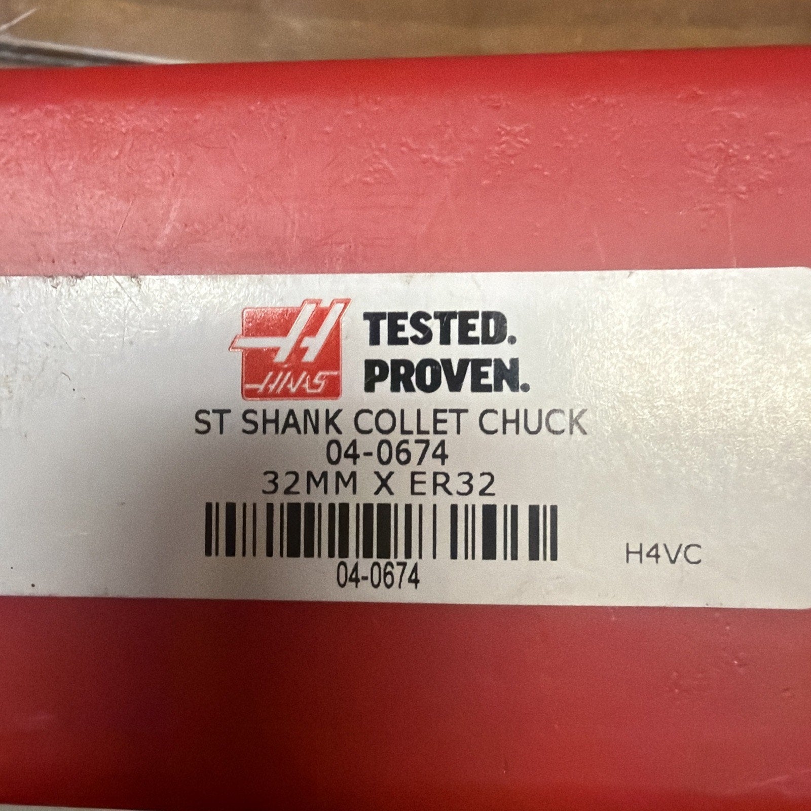 Close-up of a white label on a red surface with the Haas logo, text reading "TESTED. PROVEN. ST SHANK COLLET CHUCK 04-0674 32MM X ER32," and a barcode with the number 04-0674 beneath it.