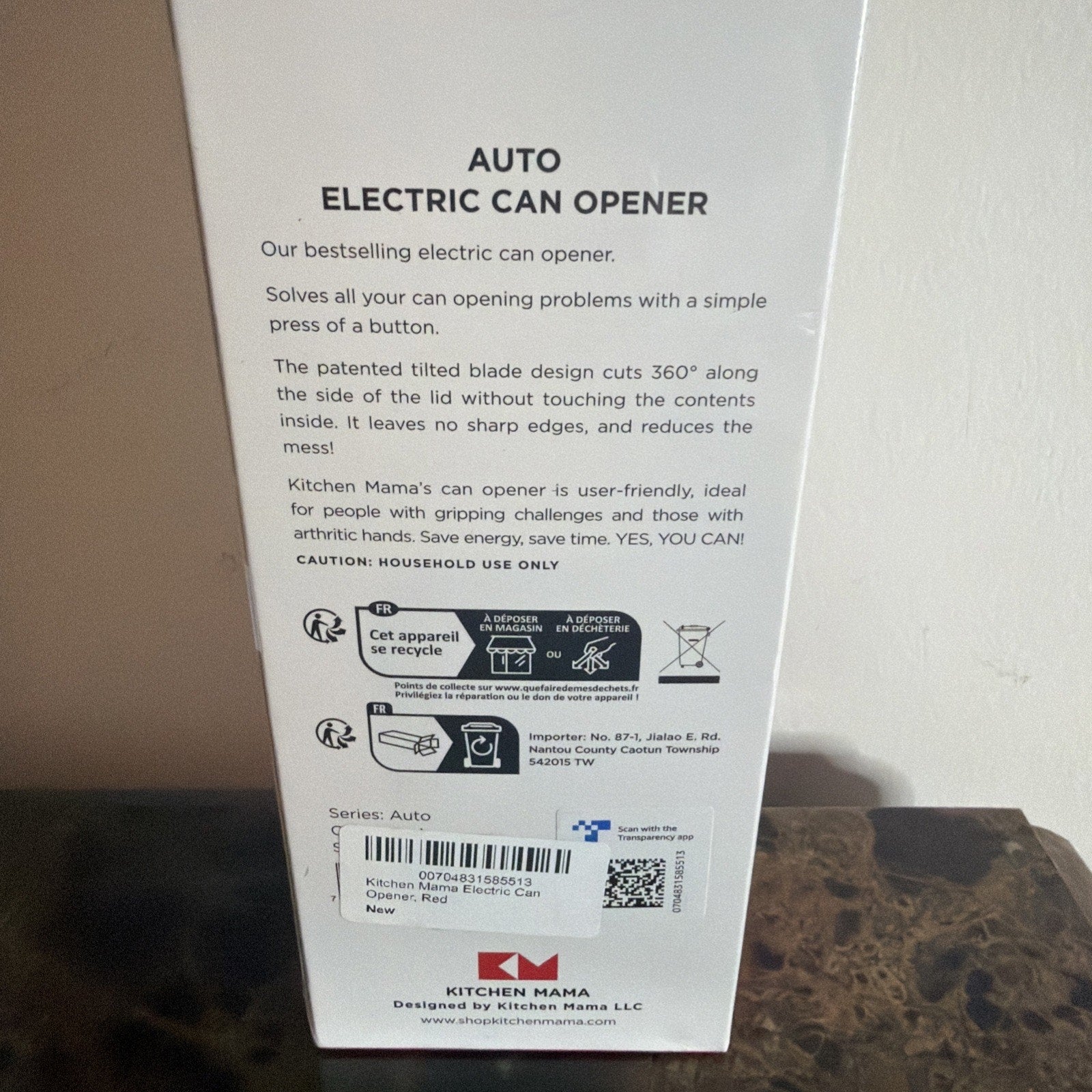 White rectangular product box standing upright on a dark marble surface, featuring printed text describing an electric can opener’s features, usage instructions, and recycling information on its back panel.