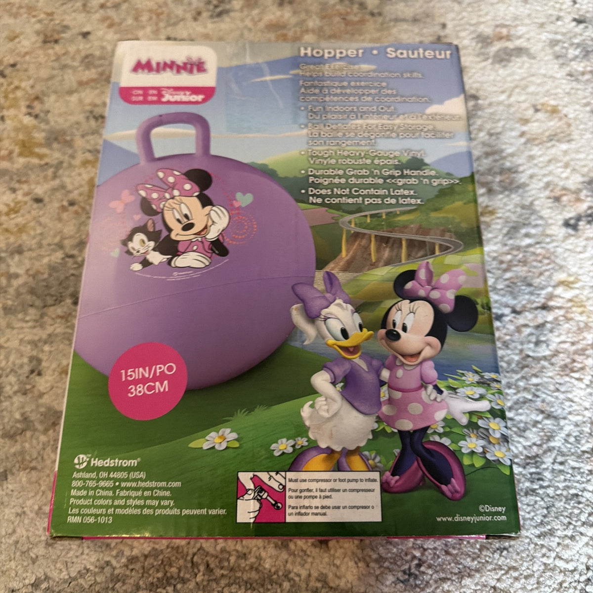 Packaging box for a purple Minnie Mouse Junior hopper ball featuring illustrations of Minnie Mouse and Daisy Duck, with text highlighting a durable grab and grip handle and size information, placed on a textured carpet.