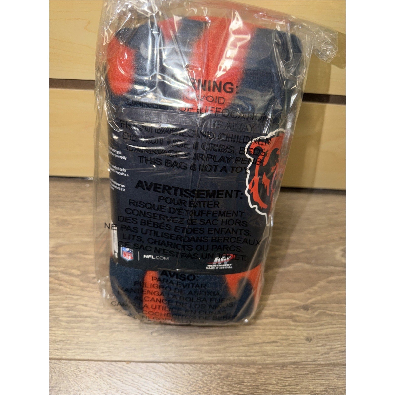 Plastic-wrapped fleece blanket featuring red and navy blue colors with a partially visible graphic logo, placed upright on a wooden floor against a light wood background.