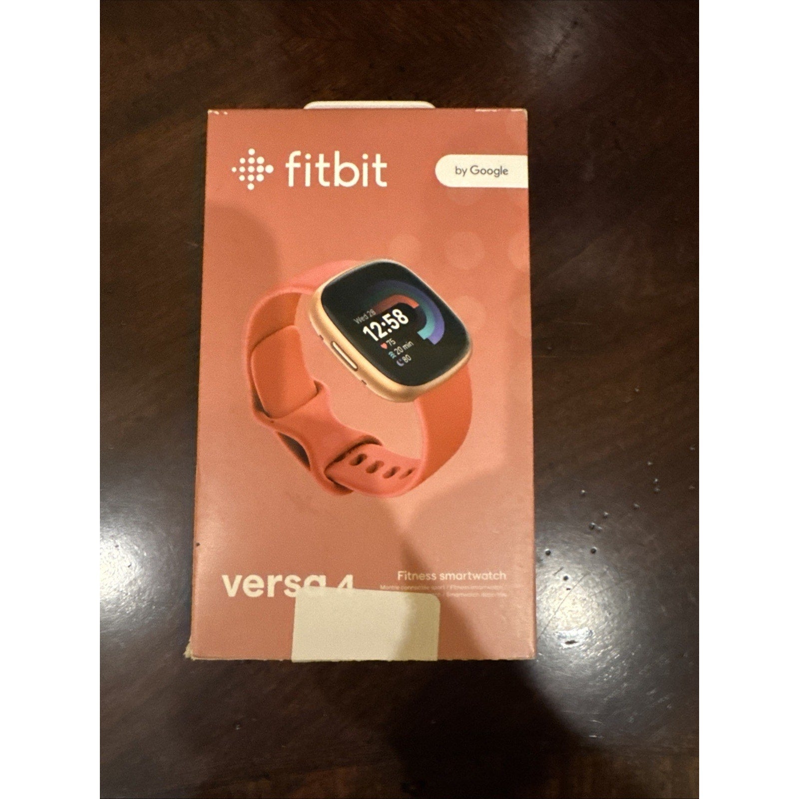 Fitbit Versa 4 fitness smartwatch in Pink Sand with Copper Rose aluminum case displayed on packaging box front