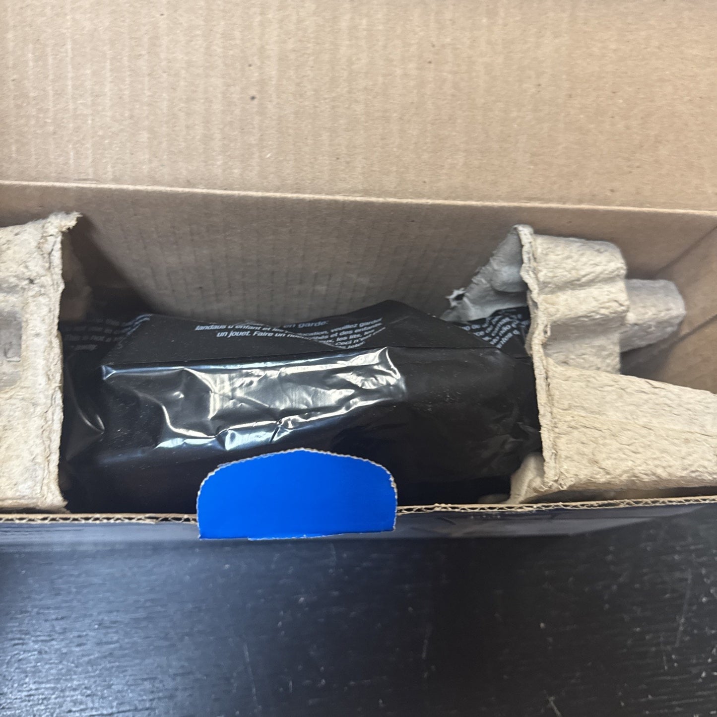 Black toner cartridge wrapped in protective plastic inside a brown cardboard box with molded pulp inserts for secure packaging on a dark surface.