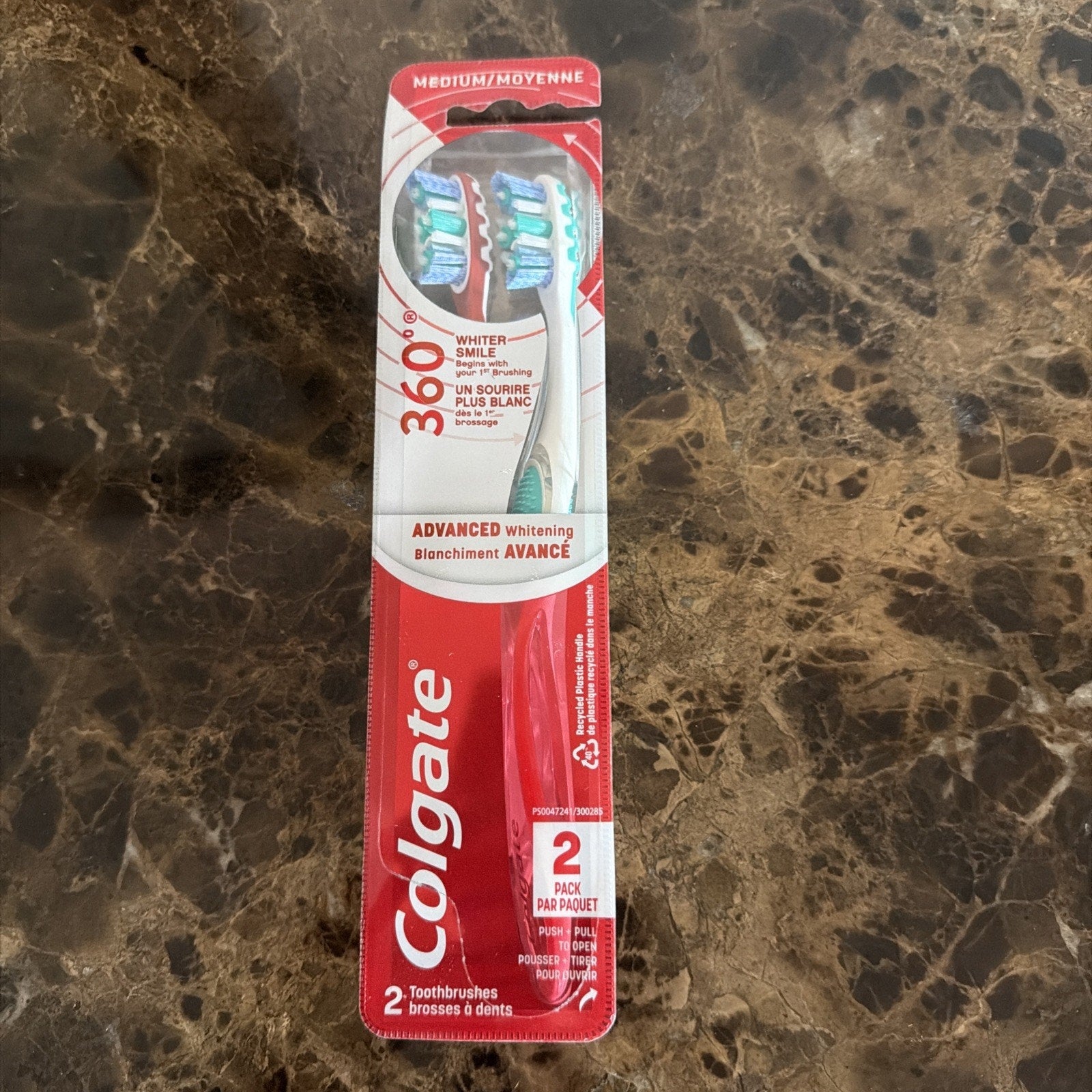 Two medium-bristle Colgate 360° Advanced Whitening toothbrushes in clear plastic packaging with red and white branding, placed on a dark marbled surface.
