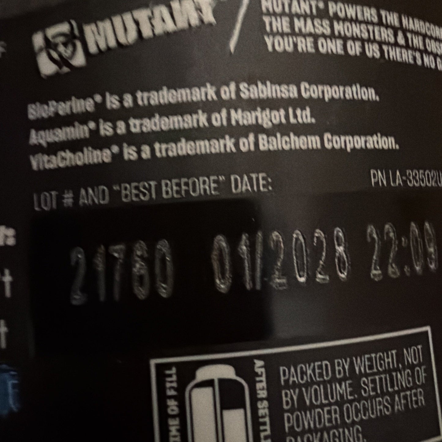 Close-up of a black supplement container showing trademark attributions for ingredients, a lot number, and a best-before date stamped as 01/2028 along with additional packaging information.