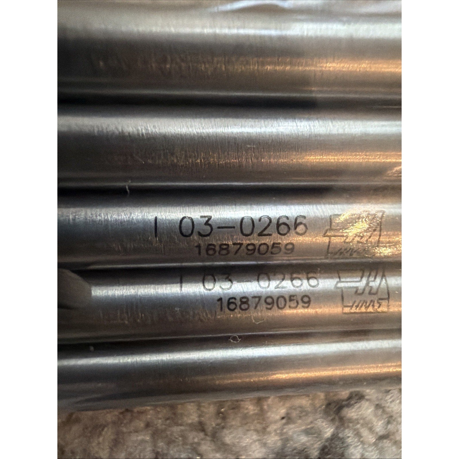 Close-up of several smooth metal cylindrical rods with engraved identification numbers and a logo, arranged horizontally on a textured beige surface.