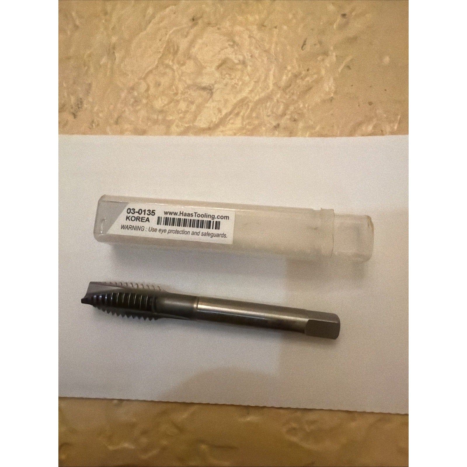 Metal threading tap with straight flutes and a hexagonal shank, placed on a white sheet next to a transparent plastic storage tube labeled with a barcode and website information.