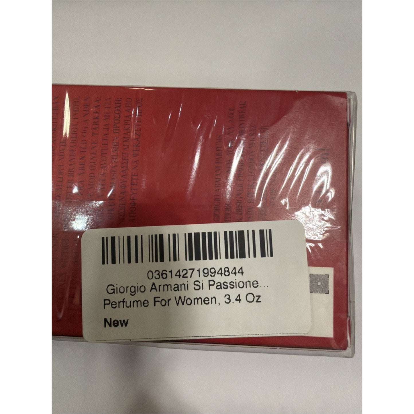 Close-up of a red rectangular perfume box wrapped in clear plastic with a white label showing barcode, product name, size 3.4 oz, and indication that it is new.
