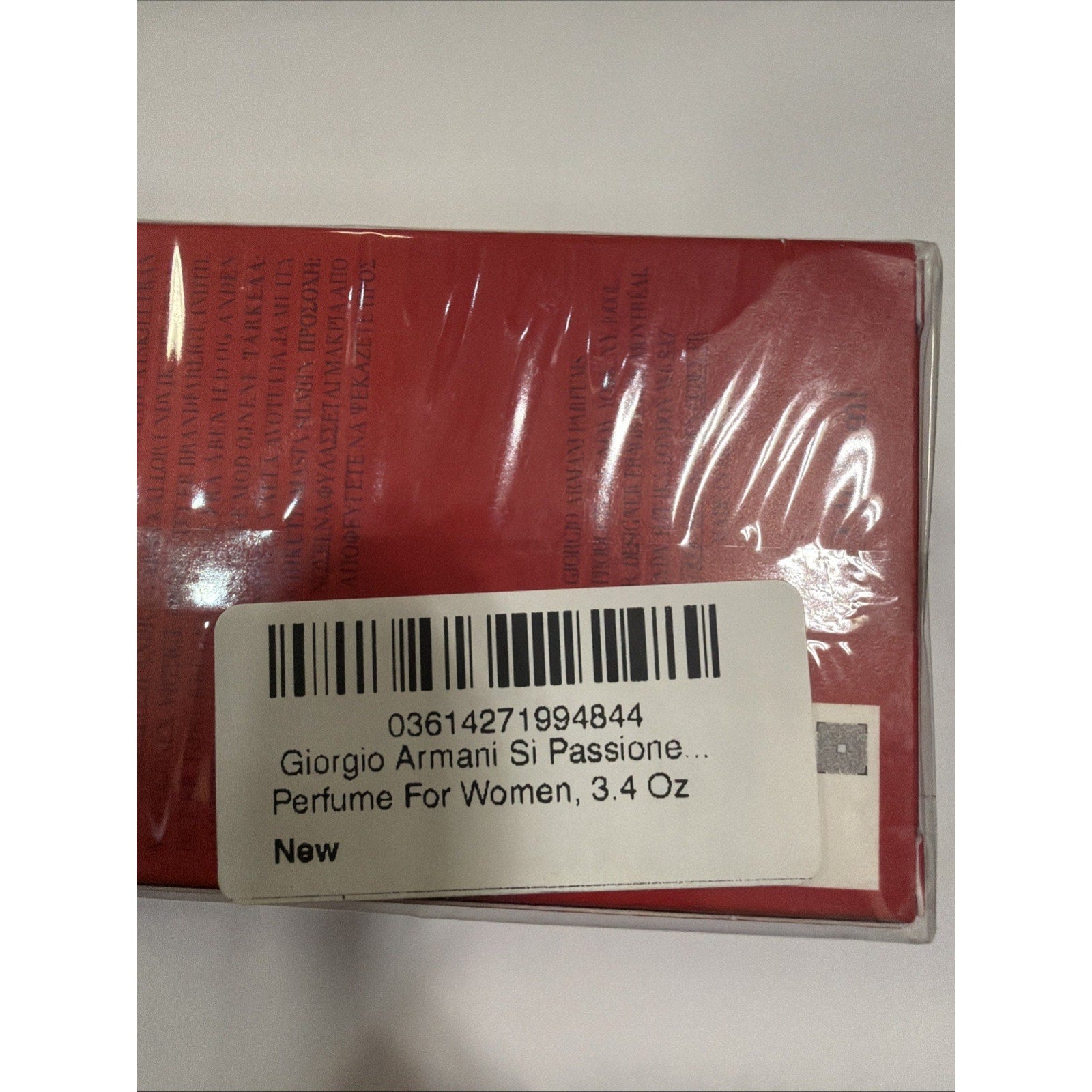 Close-up of a red rectangular perfume box wrapped in clear plastic with a white label showing barcode, product name, size 3.4 oz, and indication that it is new.