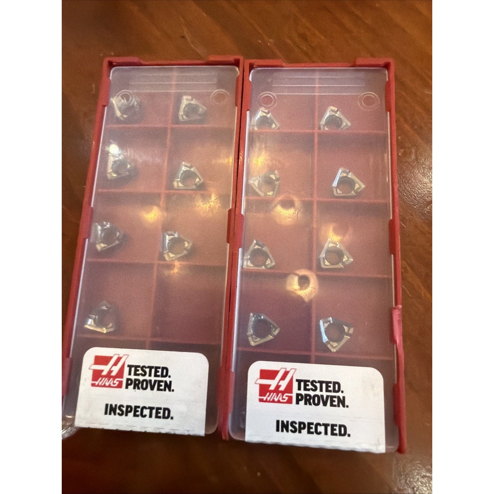 Two rectangular red plastic containers with translucent lids, each holding multiple triangular metal inserts, labeled with Haas branding and text "Tested. Proven. Inspected." on a wooden surface.