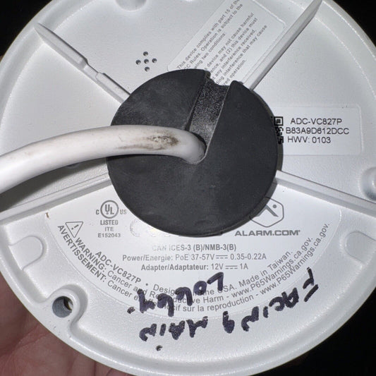 Bottom view of Alarm.com ADC-VC827P security camera showing model label, cable, and warning stickers on white casing